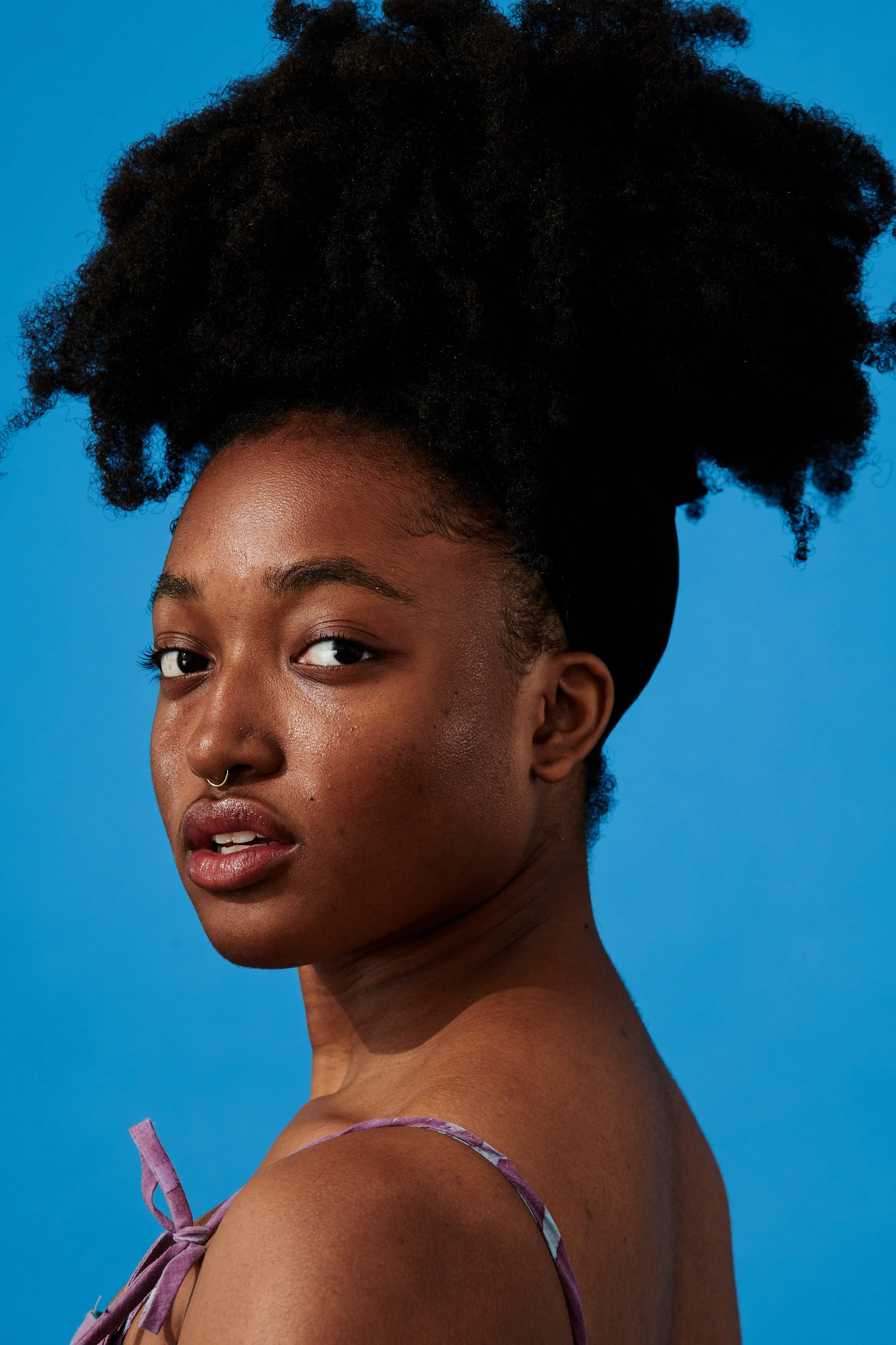 A young Black woman with natural curly hair styled up and back, wearing a purple strap top, posing against a blue background.