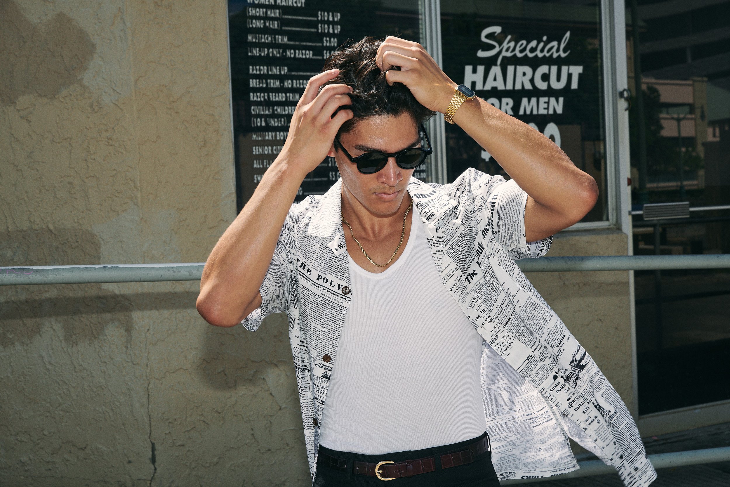 Young man with dark hair, wearing sunglasses, a newspaper-print shirt over a white t-shirt, black pants with a brown belt, standing outside a hair salon, adjusting his hair.