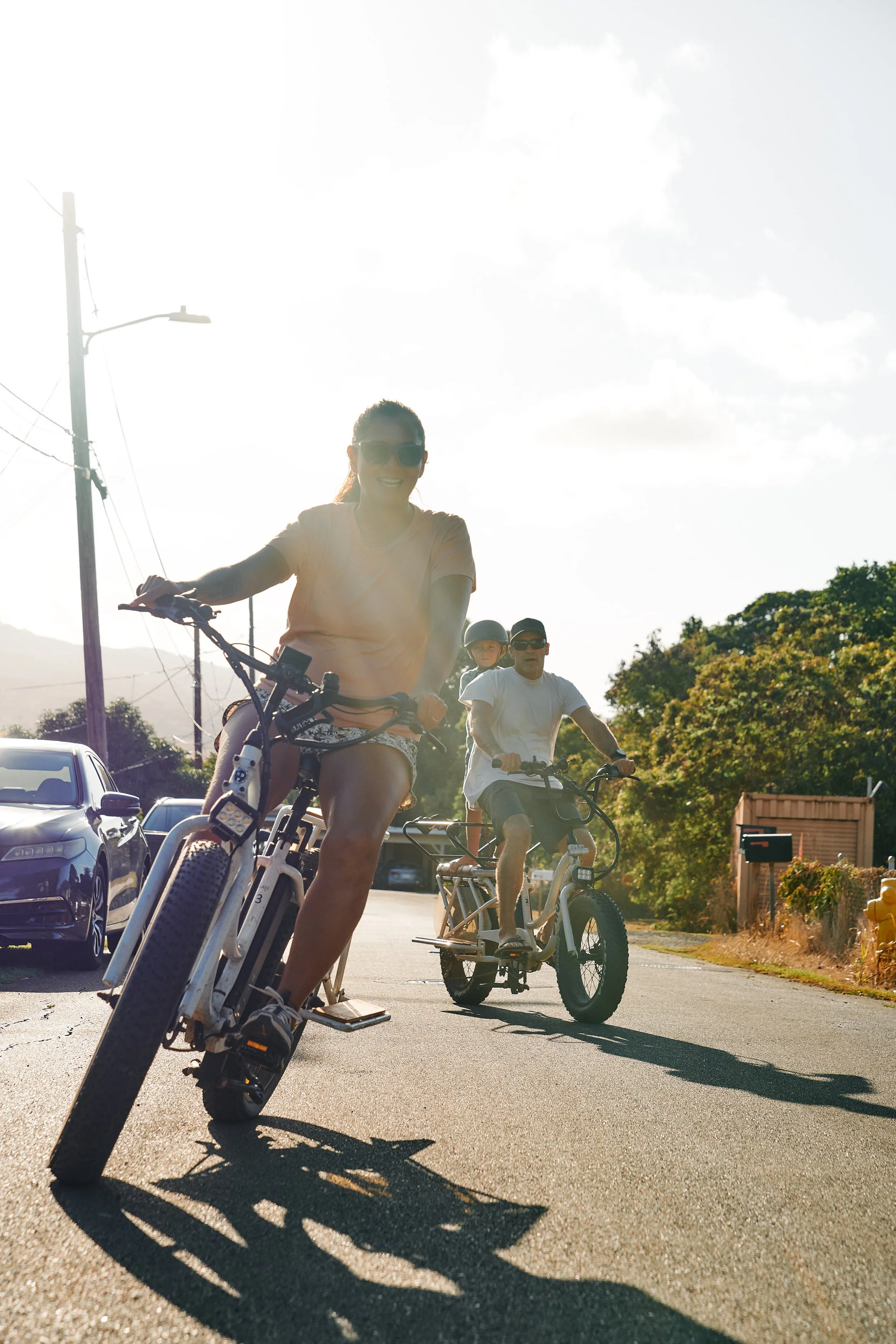 Three people riding bikes on a sunny street with parked cars and trees in the background.