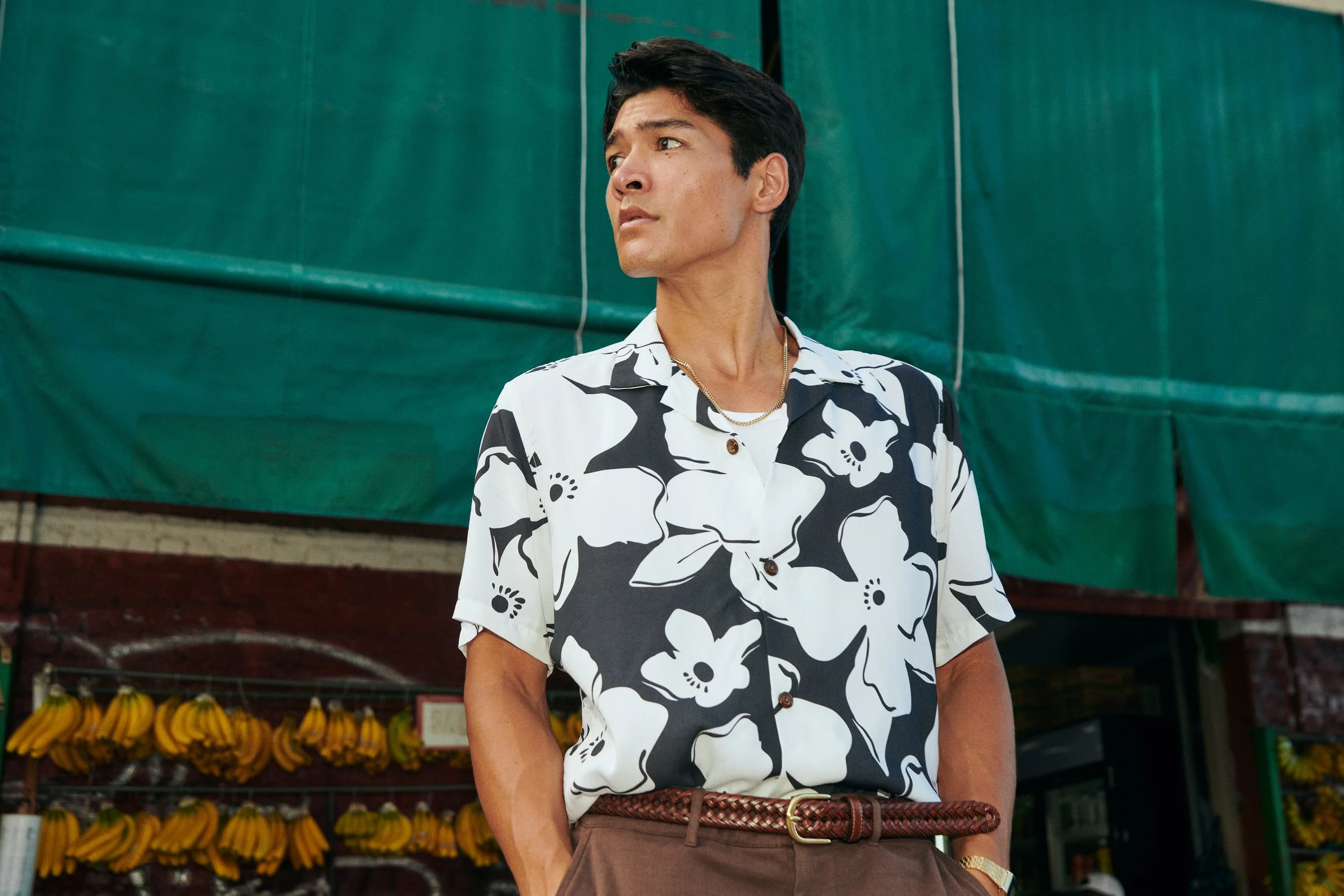 A man with dark hair and Asian features wearing a white and black floral short-sleeve shirt, brown belt, and brown pants, standing outdoors in front of a market stall with bananas hanging in the background.
