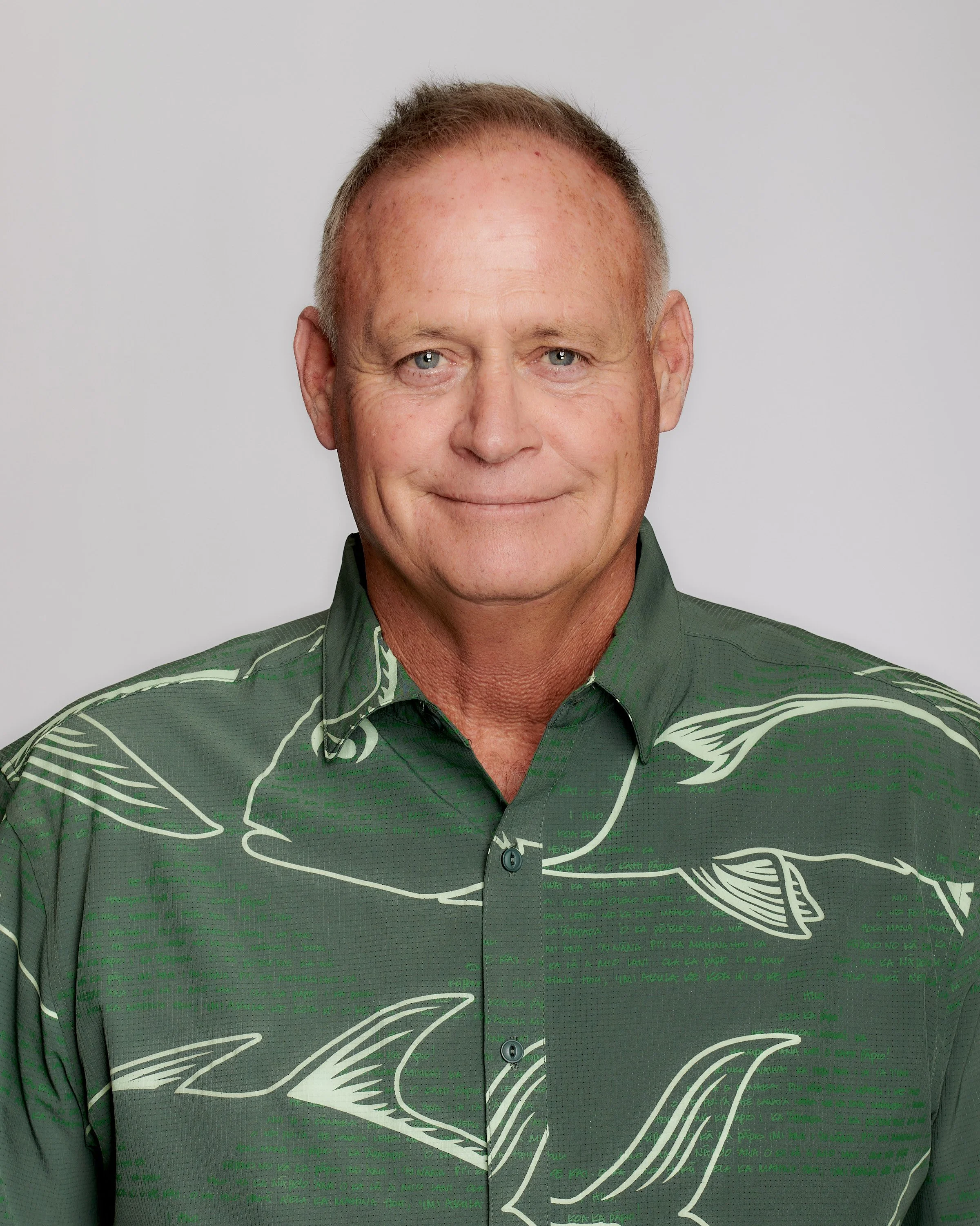 A middle-aged man with short, light-colored hair and blue eyes, wearing a green button-up shirt with a whale pattern, smiling against a plain light gray background.
