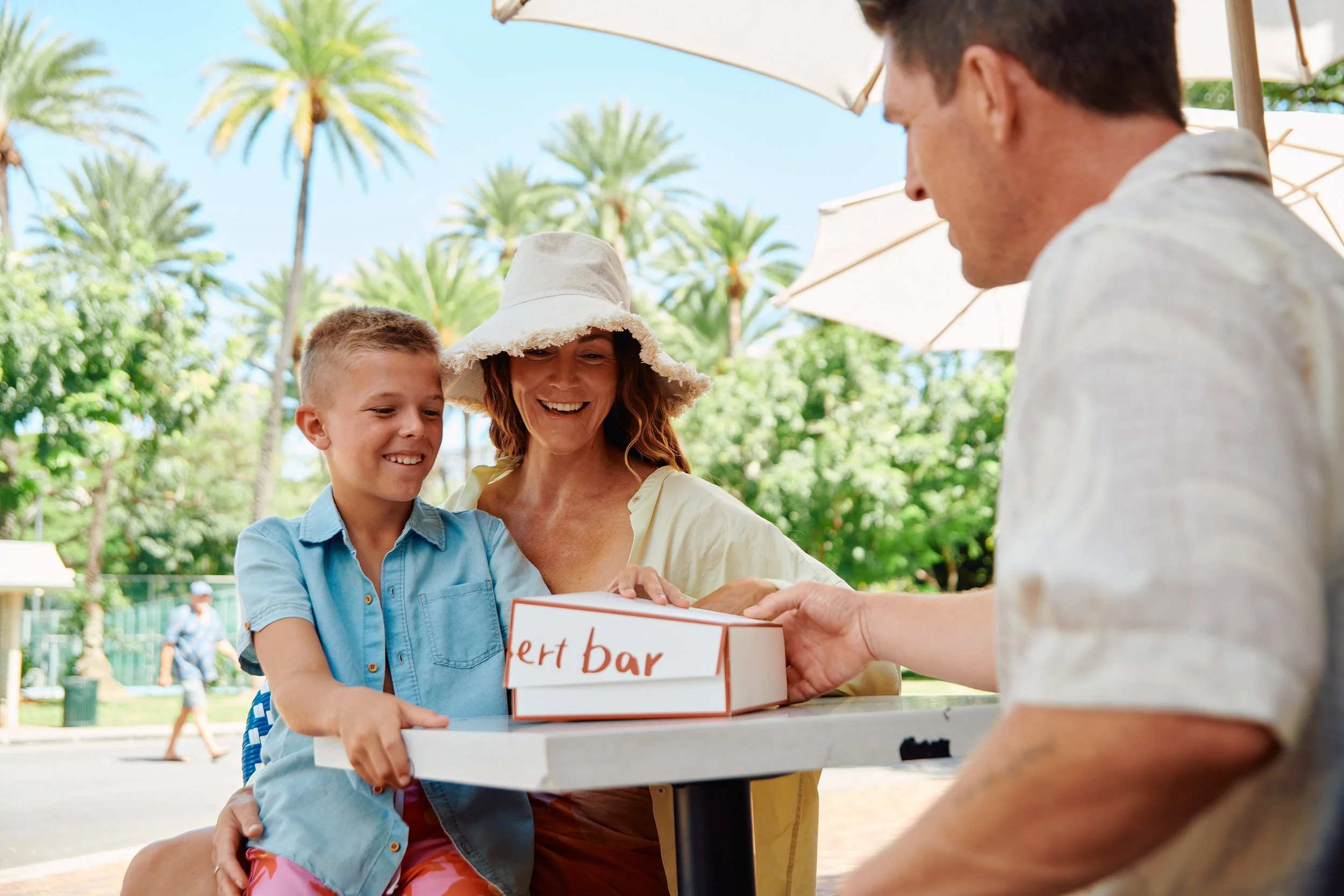A family at an outdoor cafe. A woman and young boy are smiling as they receive a pizza box from a man. The woman is wearing a wide-brimmed hat, and the boy is in a blue shirt. There are umbrellas and palm trees in the background.