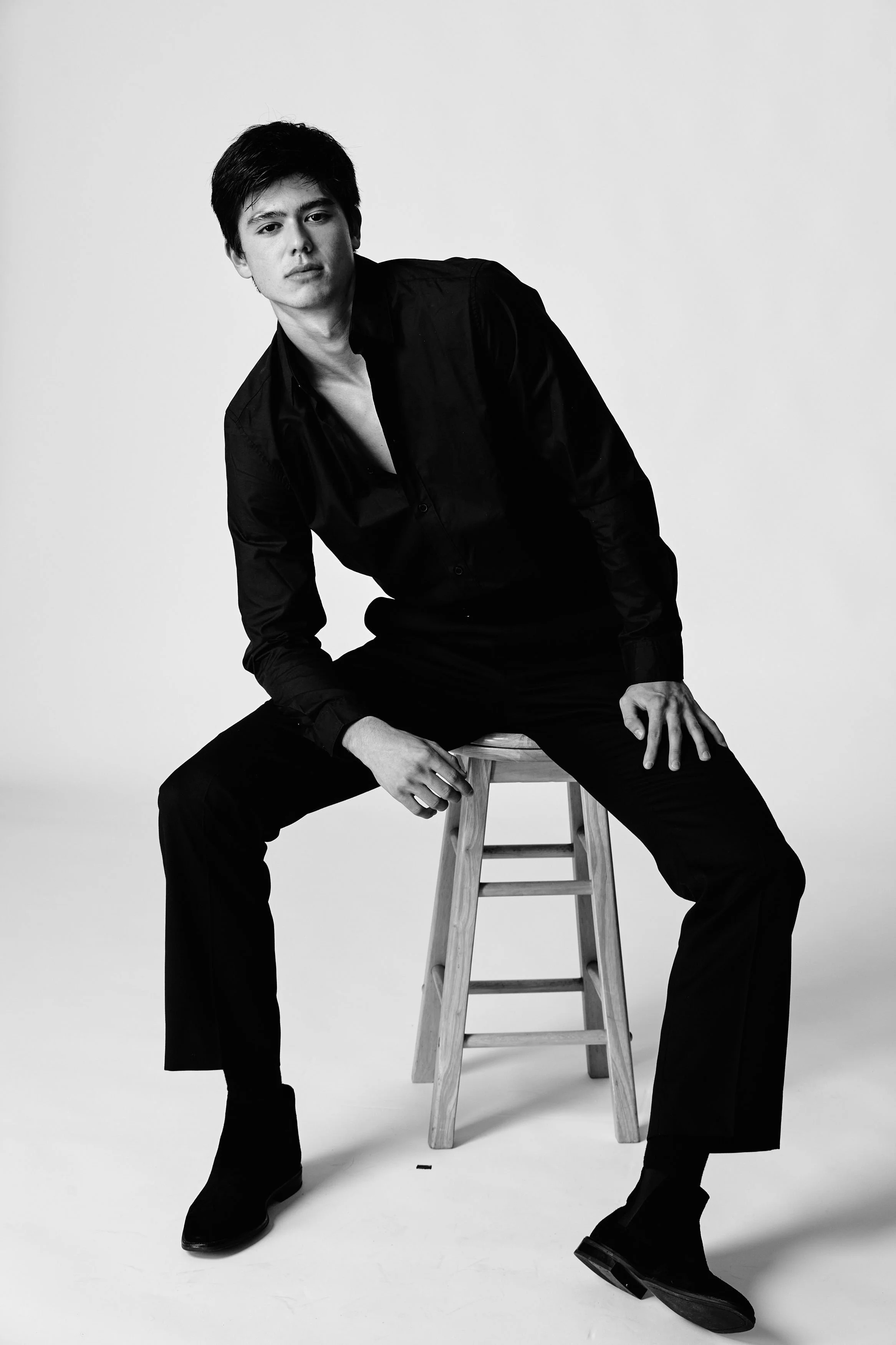 Black and white photo of a young man wearing black clothes, seated on a wooden stool, looking at the camera with a neutral expression.