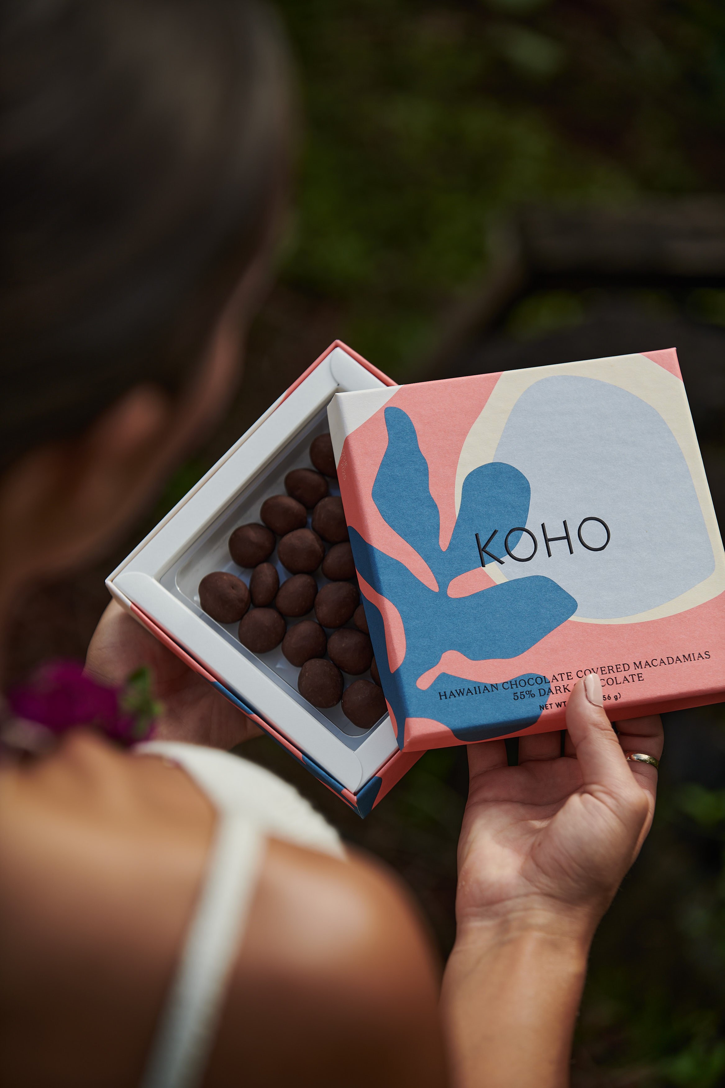 A person holding an open box of dark chocolate covered macadamia nuts from the brand KOHO. The box has a colorful abstract design.