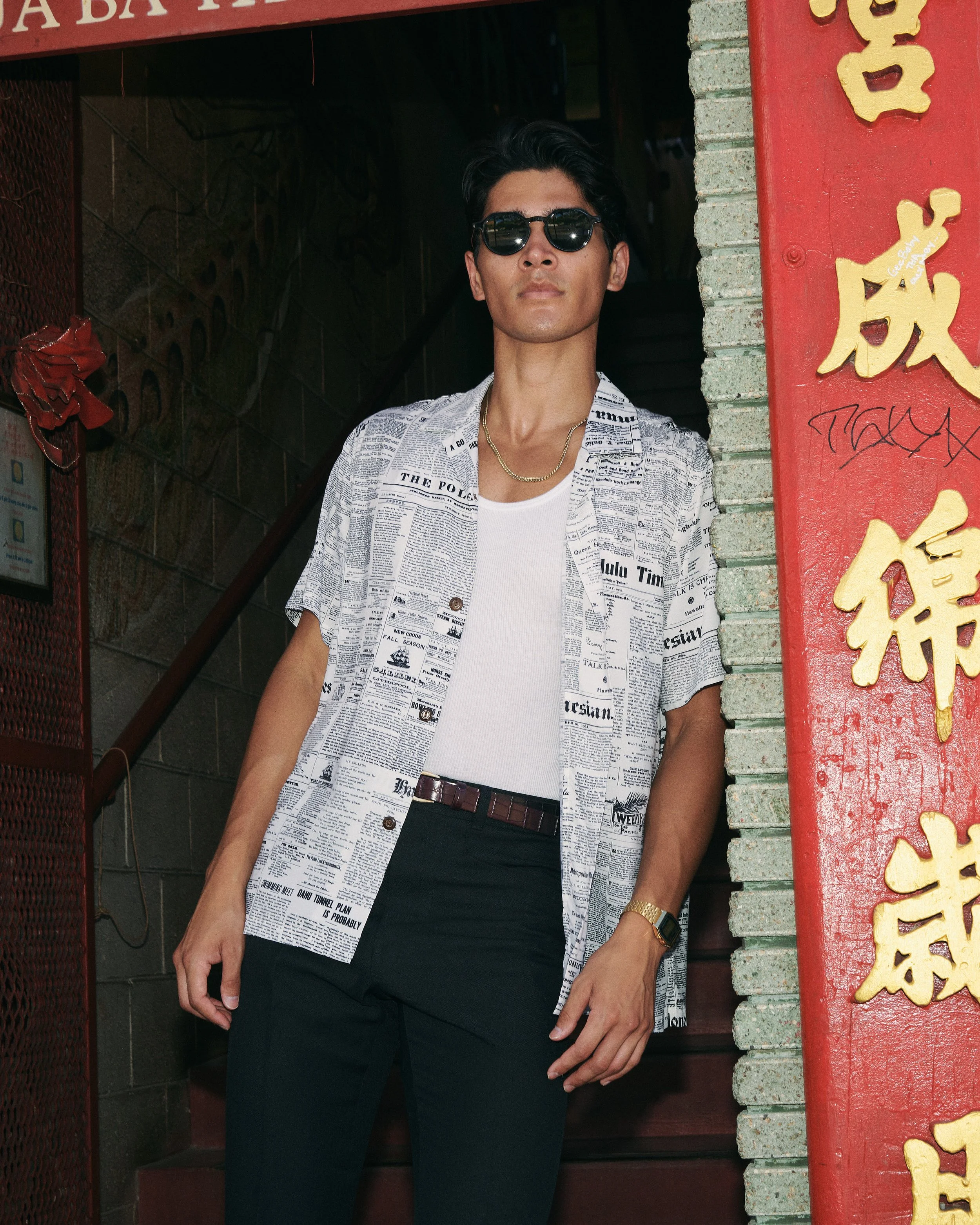 A man wearing sunglasses, a newspaper print short-sleeve shirt, a white t-shirt underneath, dark pants, a belt, a gold wristwatch, and a gold chain, standing near a red and gold Chinese signboard outside a building.