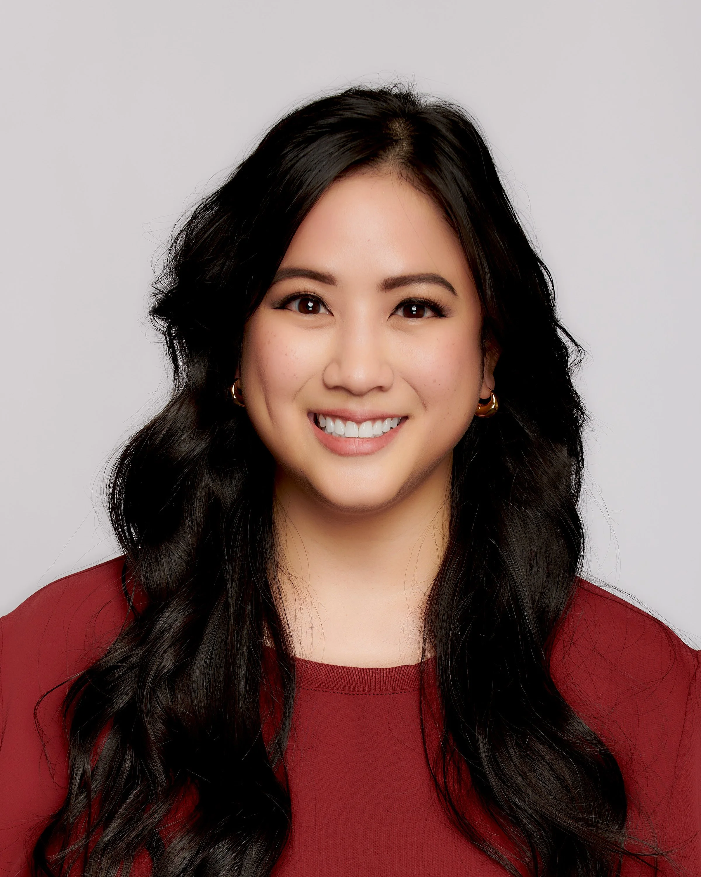 Young woman with long black hair, smiling, wearing a red top, against a plain light background.