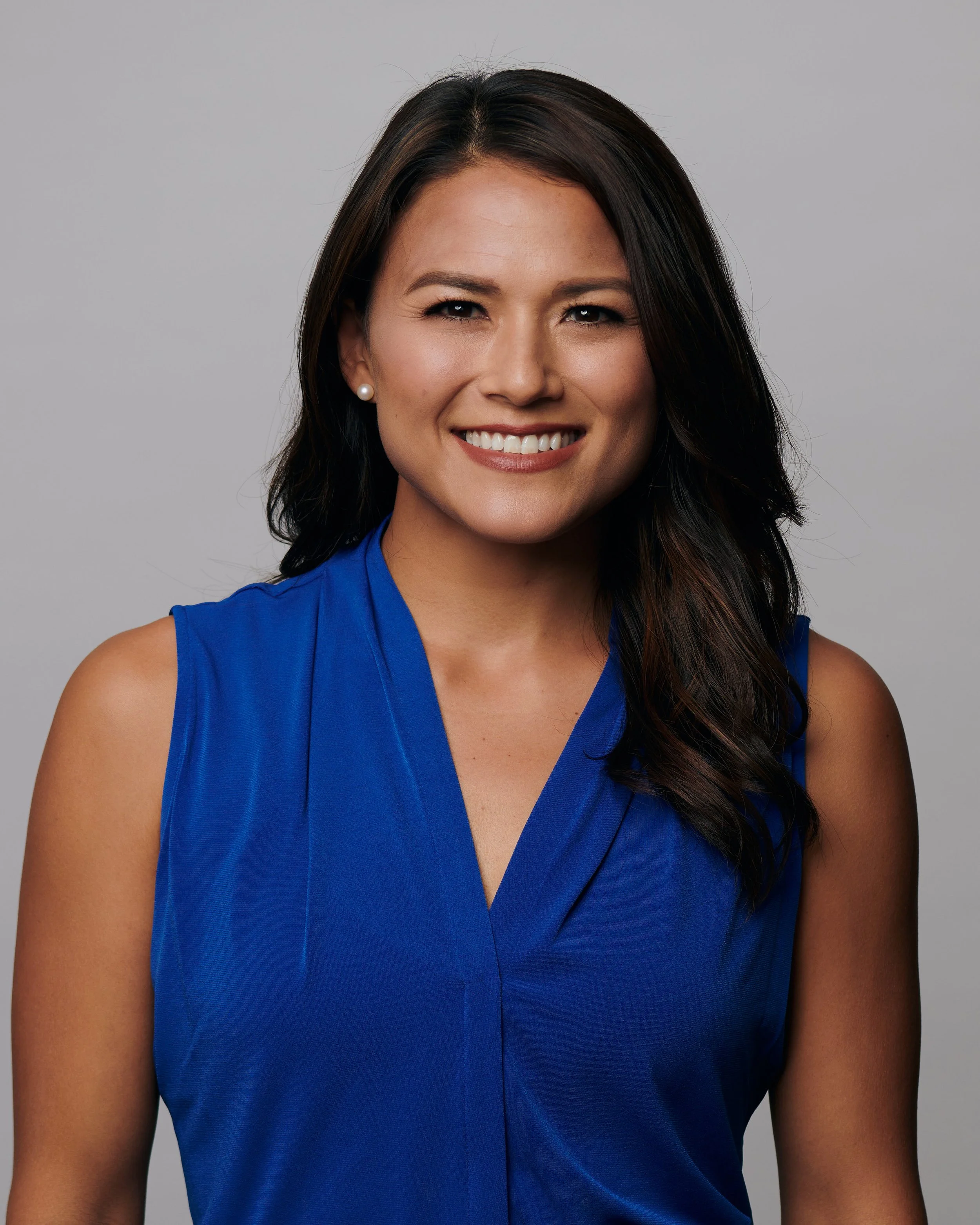 A woman with dark brown, wavy hair wearing a sleeveless blue top and pearl earrings, smiling against a plain gray background.