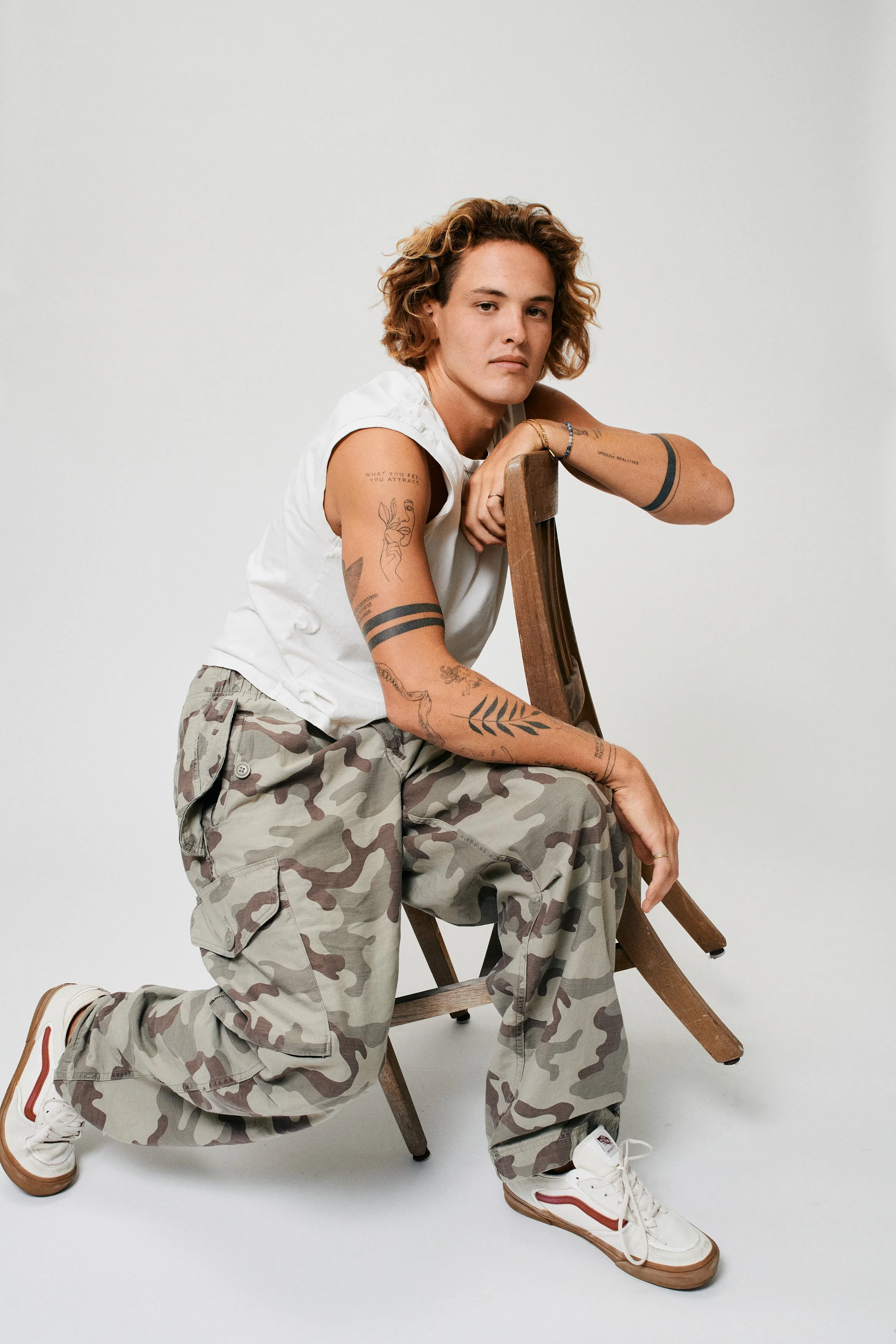 Person with tattoos wearing a white sleeveless shirt, camouflage cargo pants, and sneakers, kneeling on the floor, leaning on a wooden chair with a plain light background.