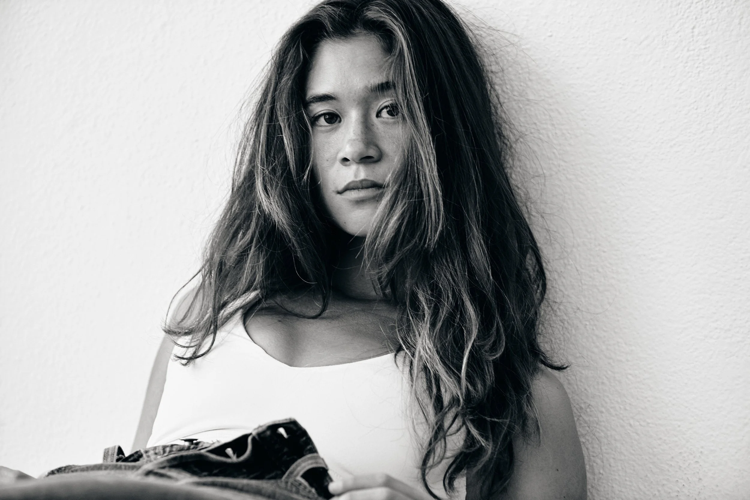 Black and white portrait of a young woman with long, wavy hair, wearing a sleeveless top, holding a pair of shoes, standing against a plain wall, looking at the camera.