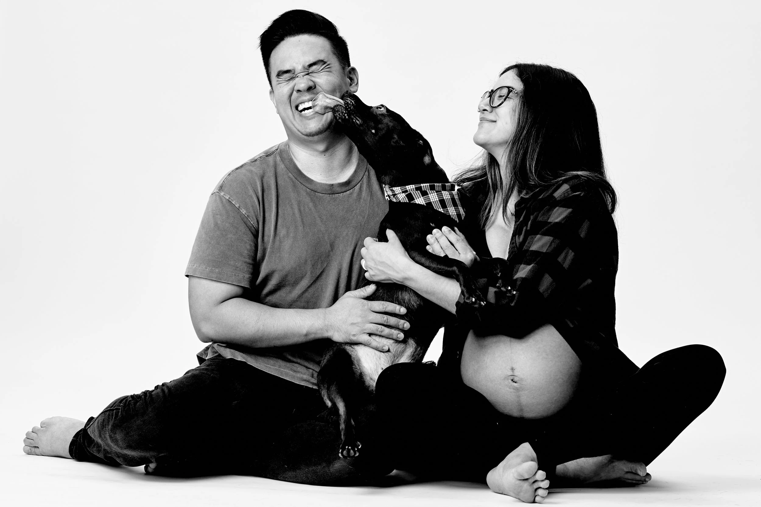 A pregnant woman sitting cross-legged and a man sitting beside her with a dog between them. The dog is licking the man's face, and both seem happy in a joyful moment.