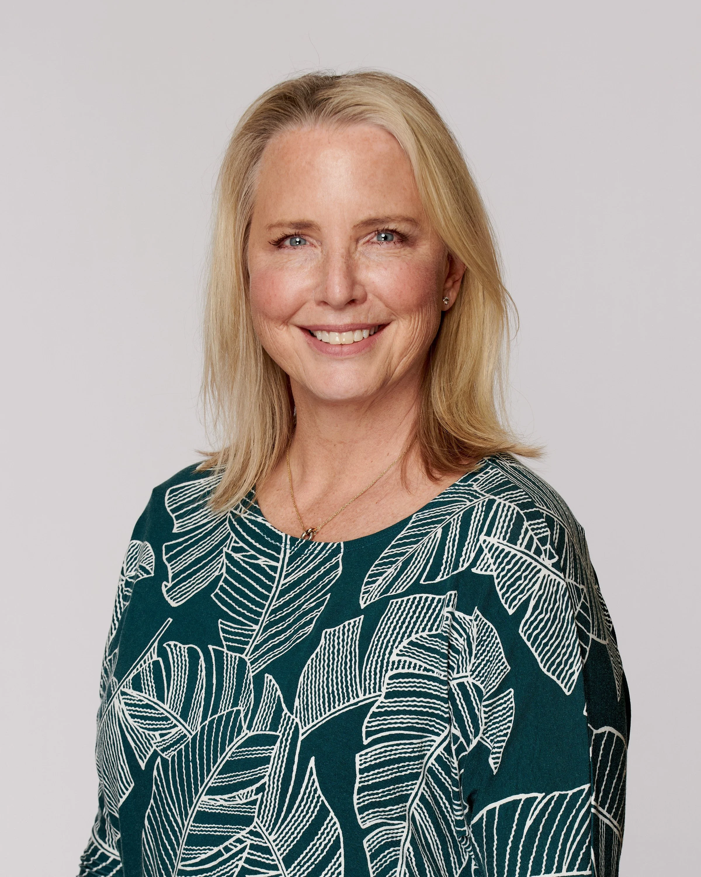 A smiling woman with blonde hair wearing a dark green top with white leaf patterns, standing against a plain light background.