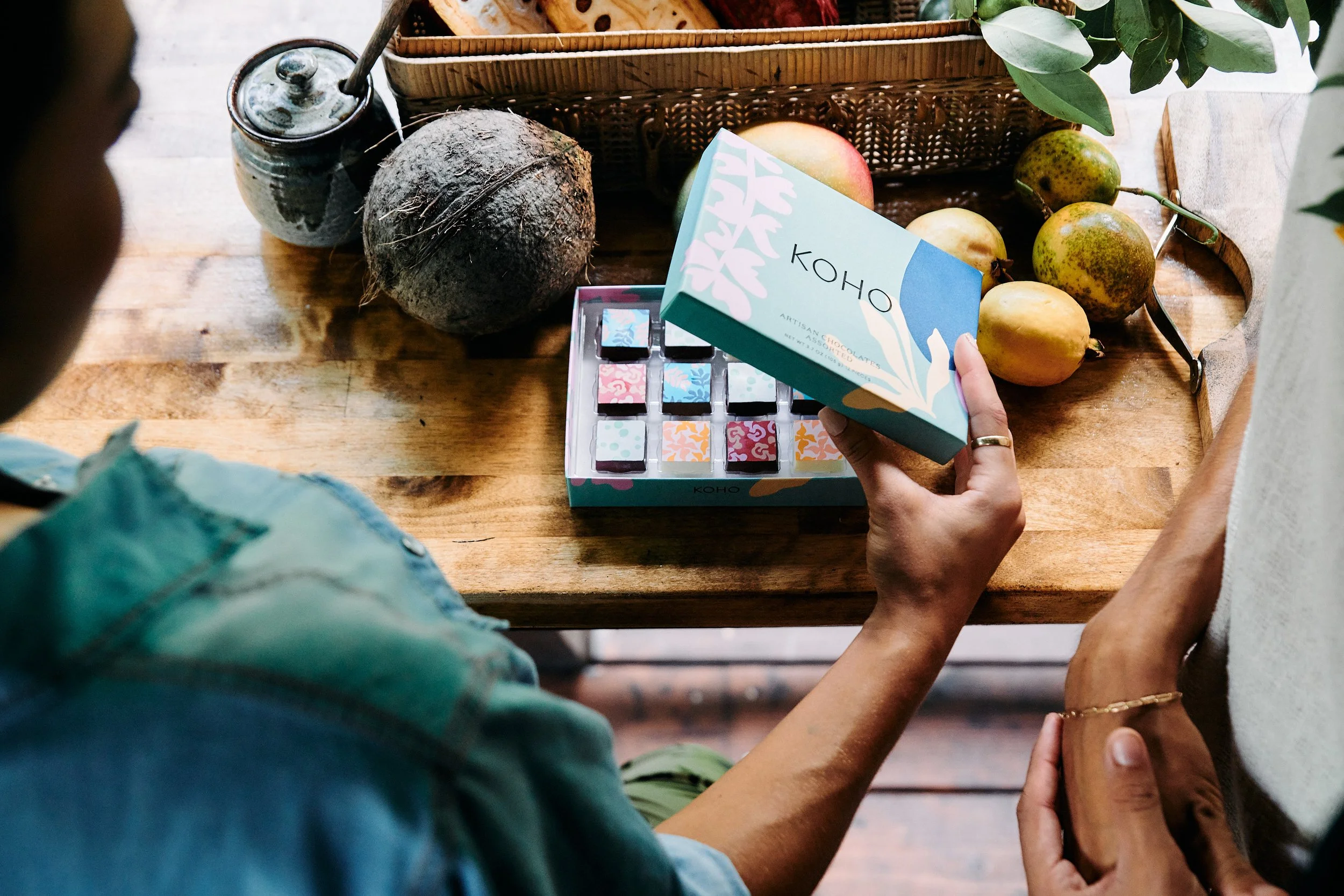 A person holding a box of KOHO artisan chocolates with colorful patterned chocolates inside, on a wooden table with tropical fruits, a coconut, a jar, and green leafy plants nearby.