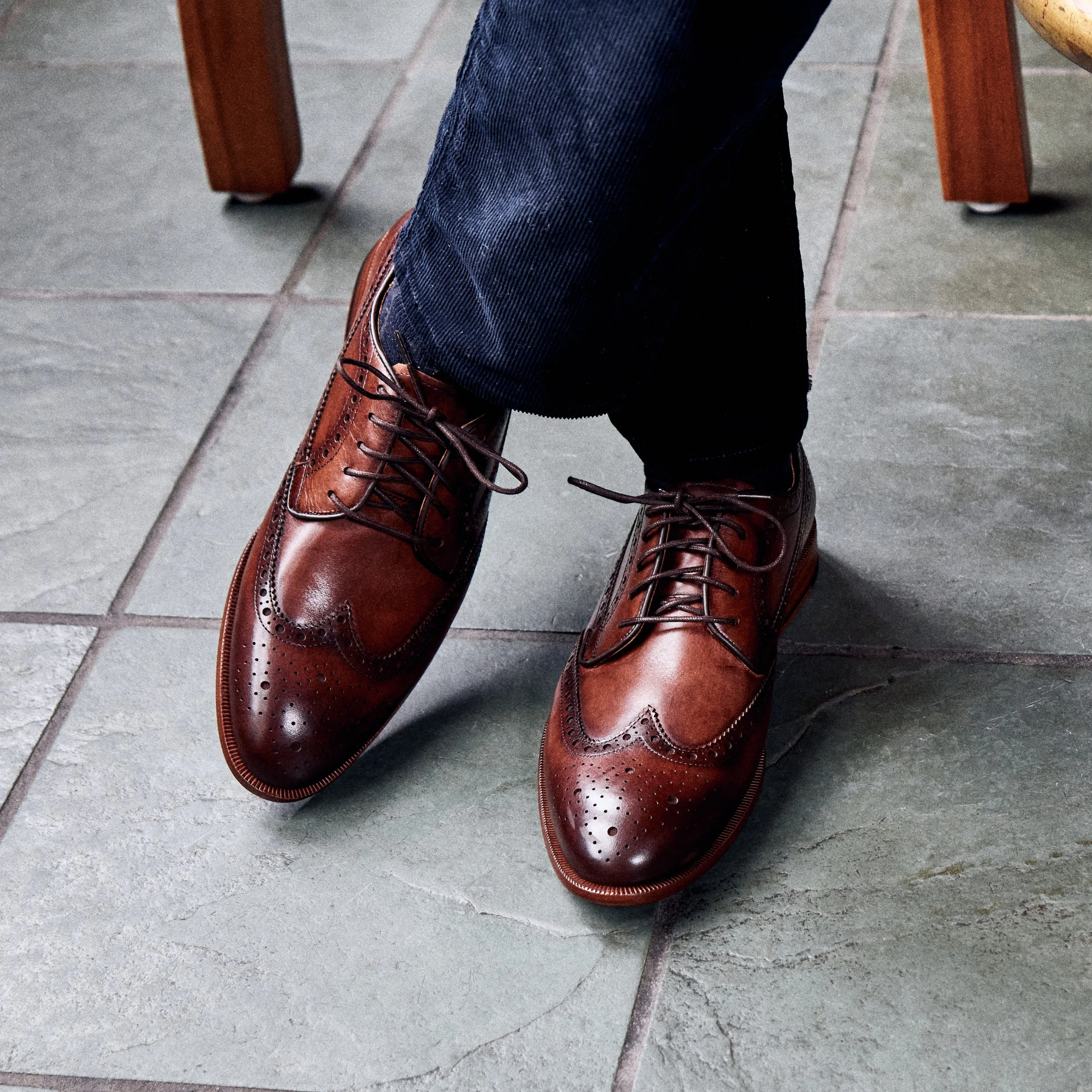 Close-up of a person wearing brown leather brogue shoes and dark blue jeans, sitting on a tiled floor.