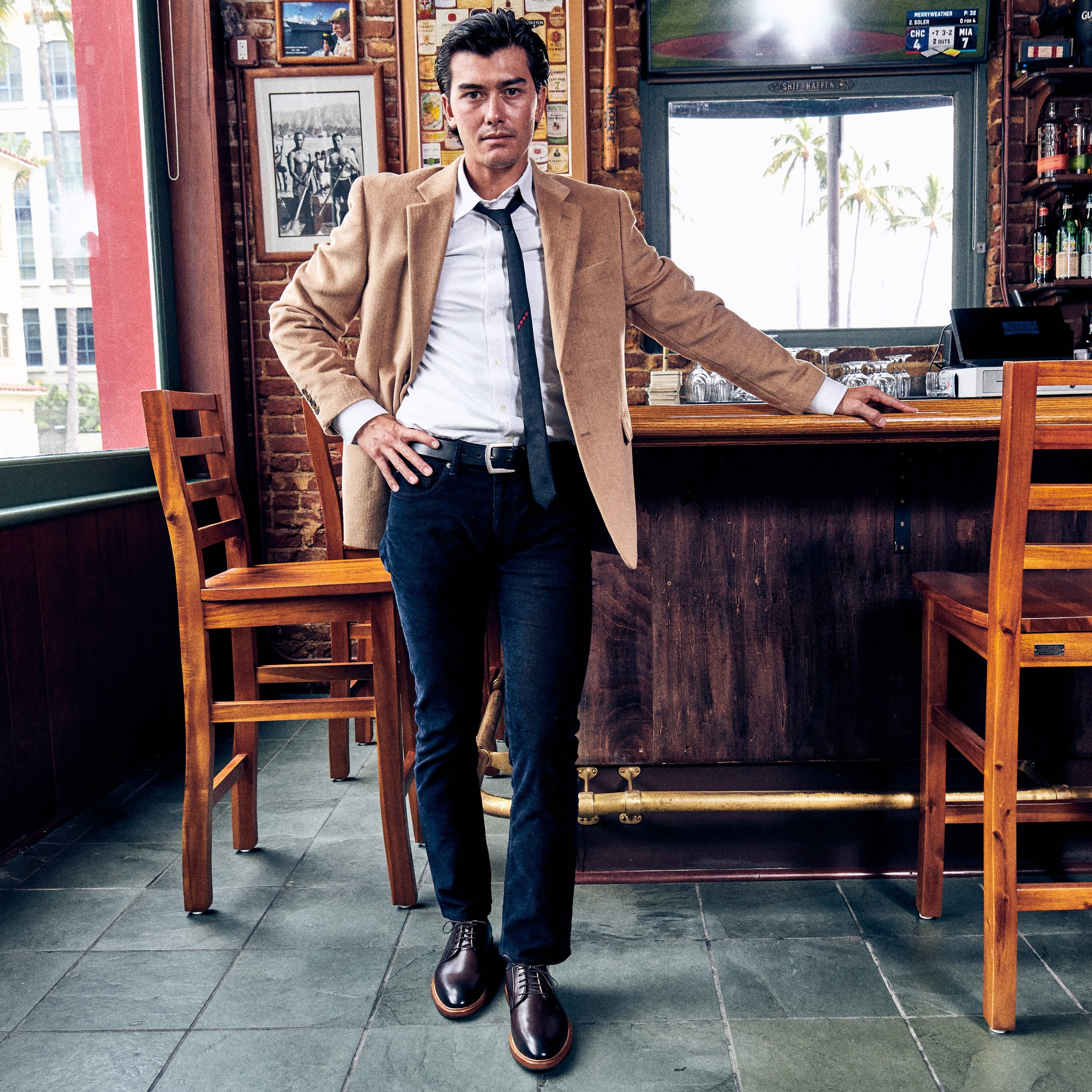 A man in a beige blazer, white shirt, black tie, and black jeans stands in a bar, leaning on the counter with his right hand. He has dark, wavy hair and a serious expression. The bar has wooden chairs and a brick wall with framed pictures, and there 