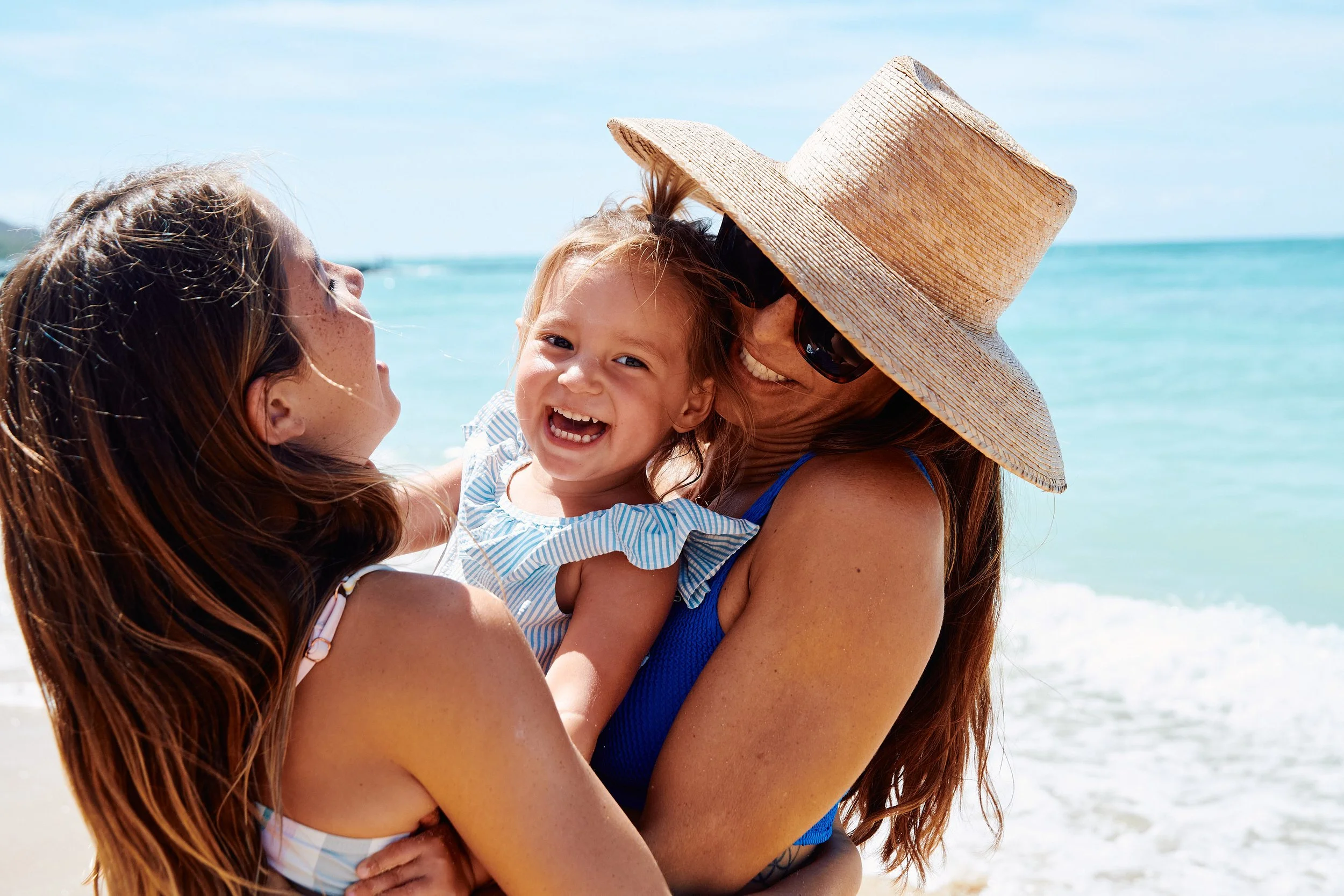 Three women and a young girl enjoying a day at the beach, smiling and hugging each other near the ocean with clear blue sky.