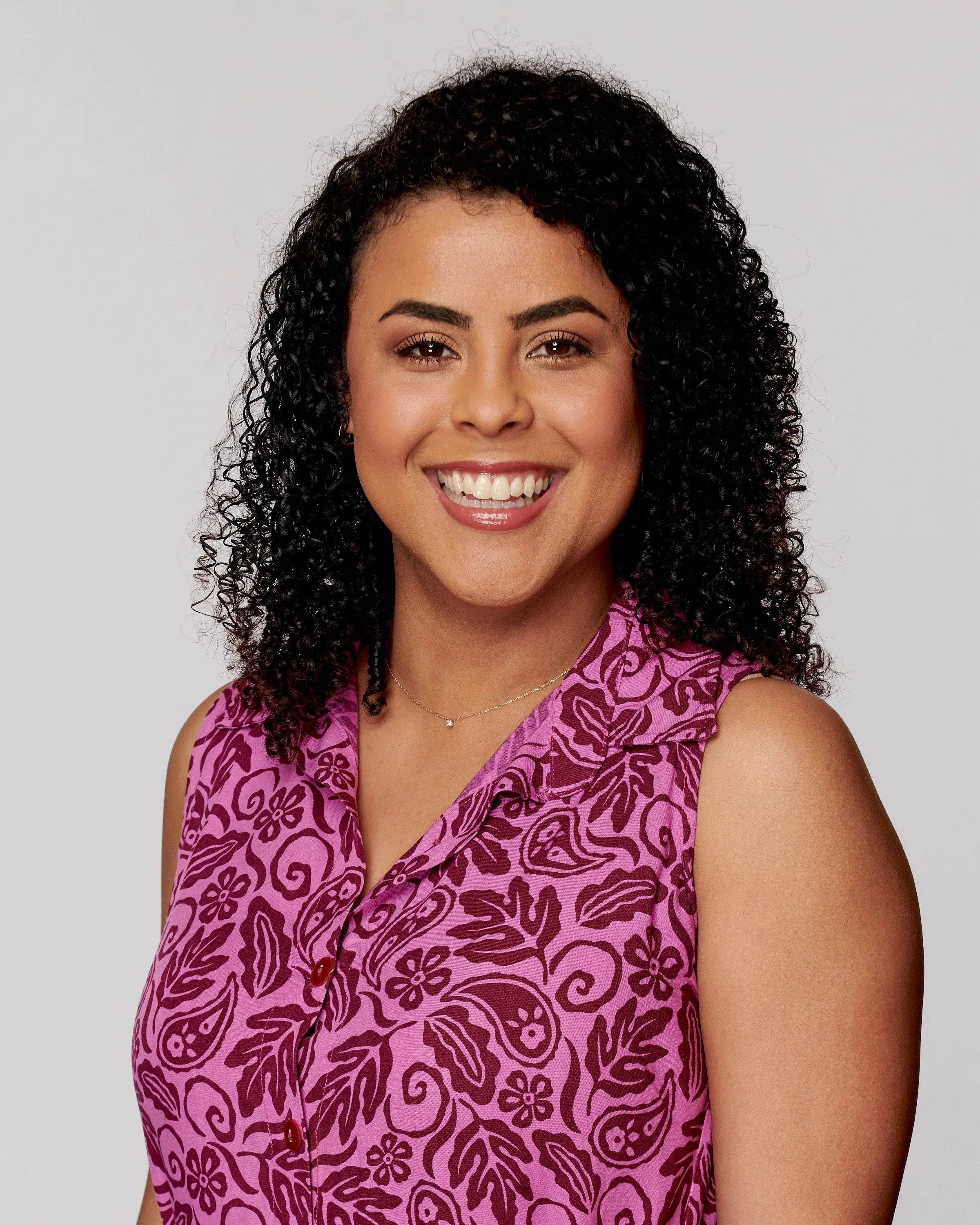 Portrait of a woman with curly black hair wearing a sleeveless pink blouse with purple floral and swirl patterns, smiling against a plain white background.