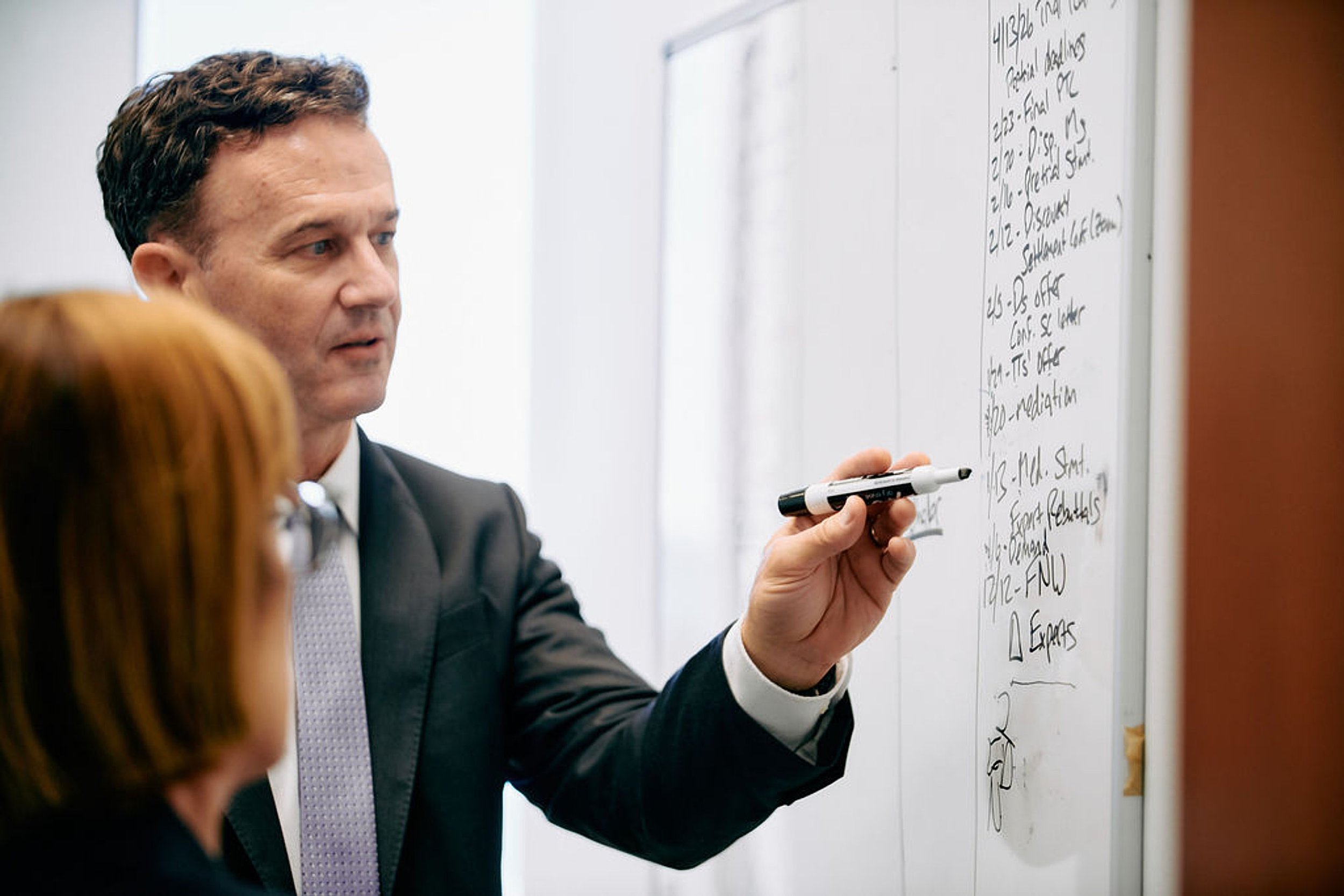 A man in a business suit writing on a whiteboard with a black marker, while a woman with red hair listens.