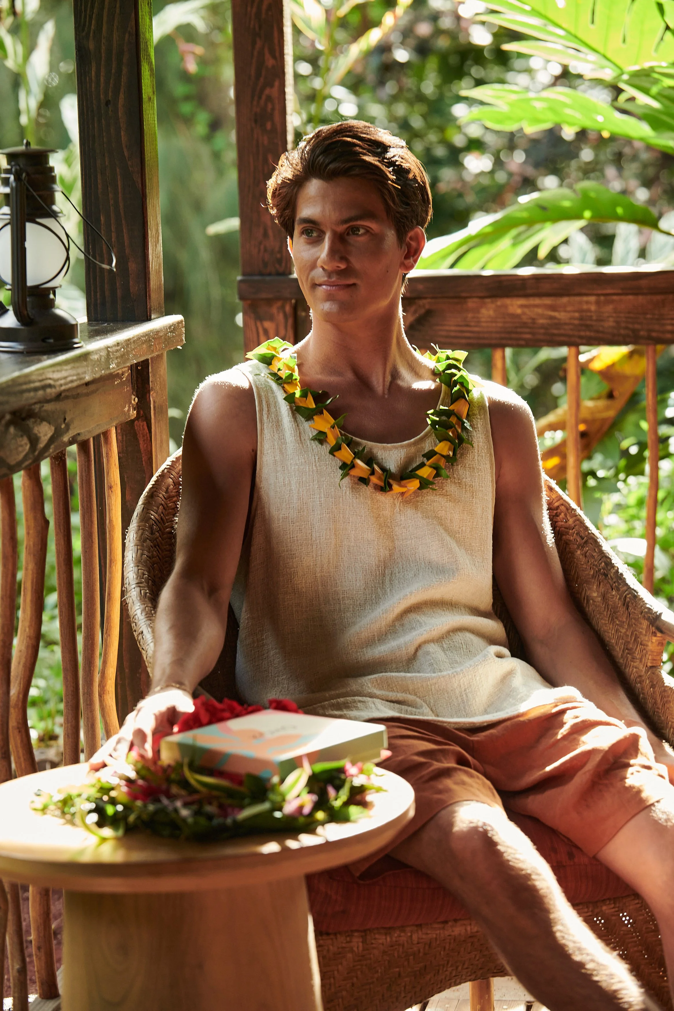 A young man sitting in a wicker chair on a wooden porch, wearing a sleeveless beige shirt and orange shorts. He has a leafy floral necklace and is surrounded by lush greenery. There is a small wooden table in front of him with a gift box, flower garl