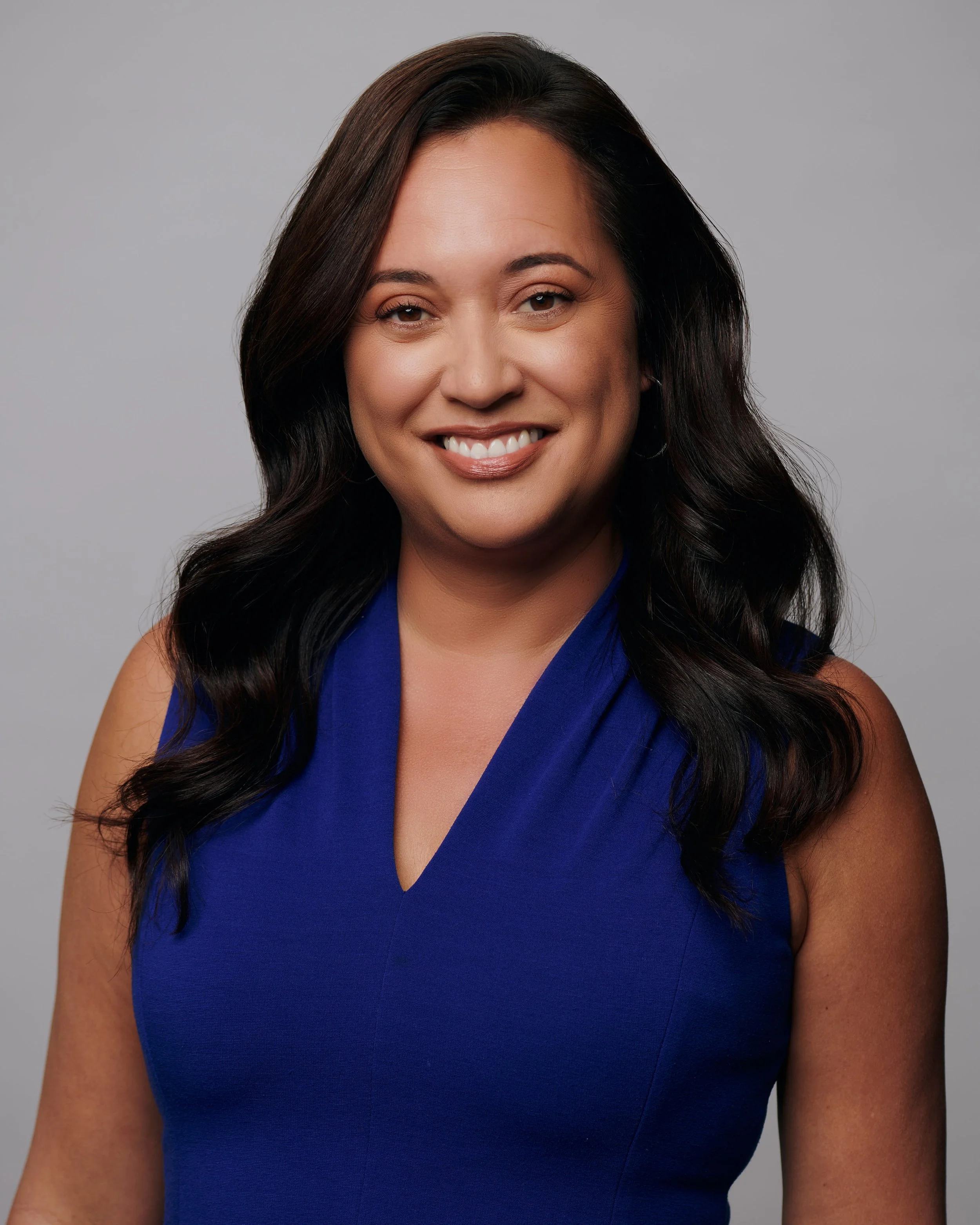 Portrait of a woman with long dark wavy hair, smiling, wearing a sleeveless royal blue top, against a plain light grey background.