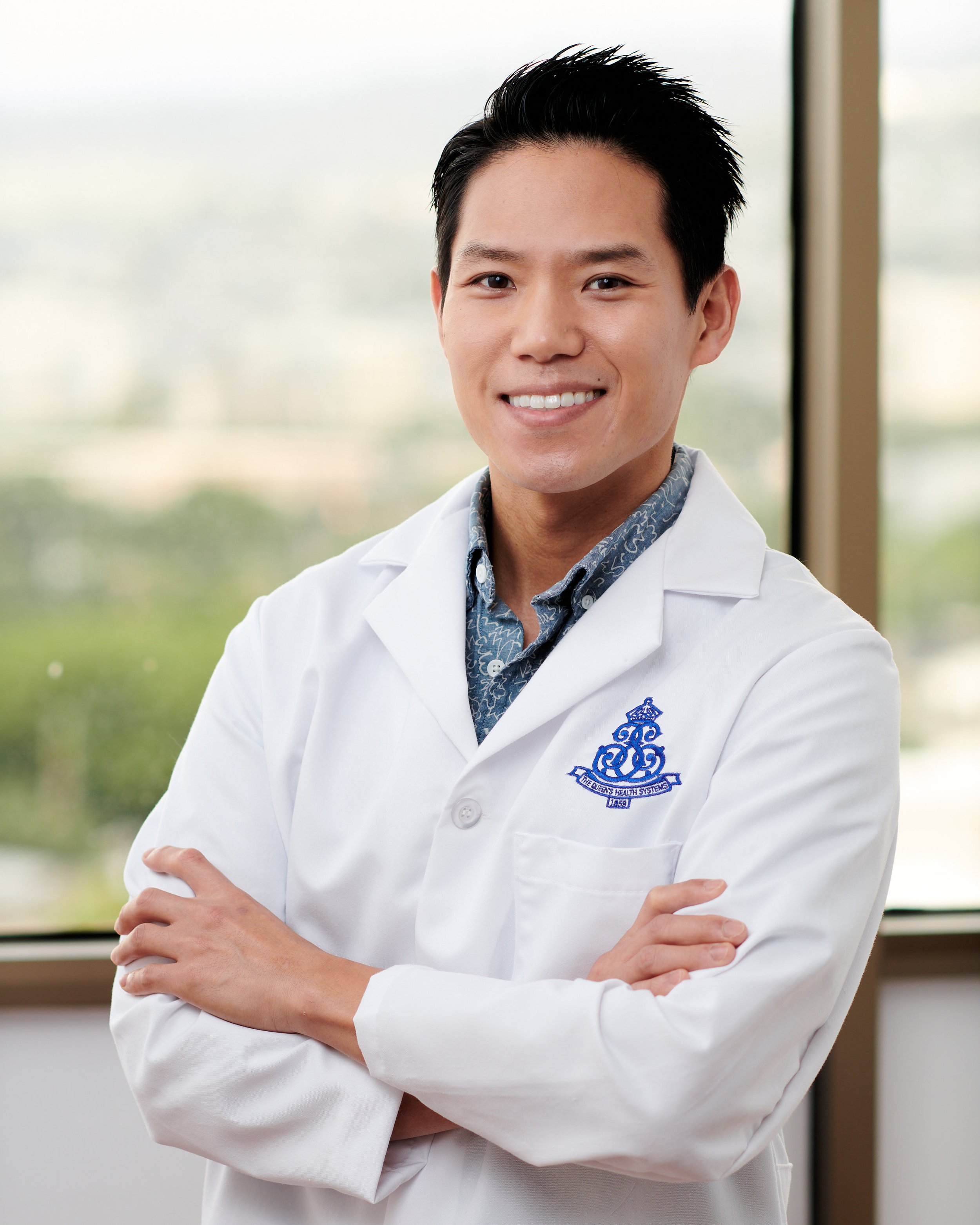 Smiling male doctor with crossed arms wearing a white lab coat and patterned shirt in front of a window with a view outside.