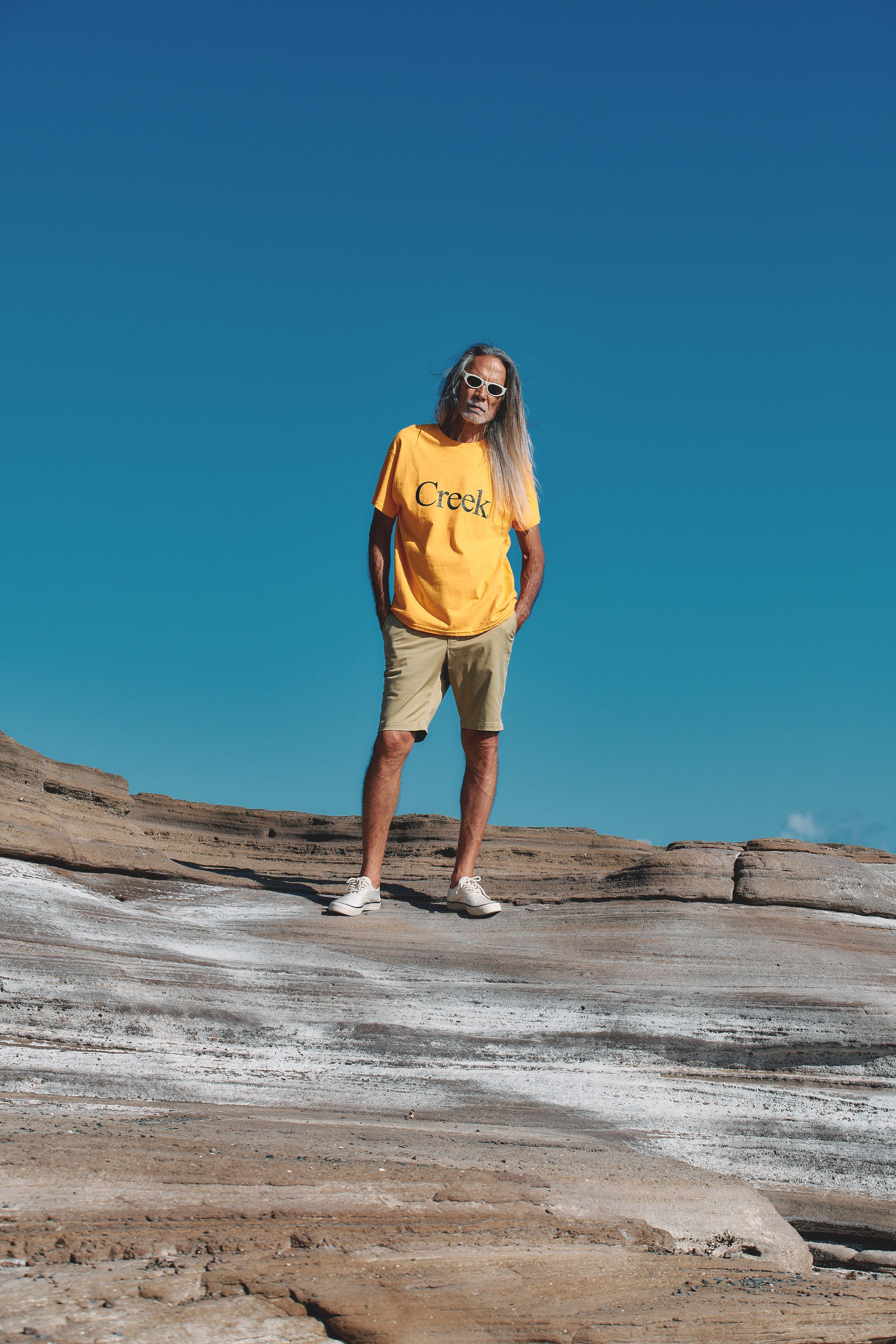 An older man with long gray hair wearing sunglasses, a yellow t-shirt with 'Creek' printed on it, beige shorts, and white sneakers standing on layered rock formations under a clear blue sky.