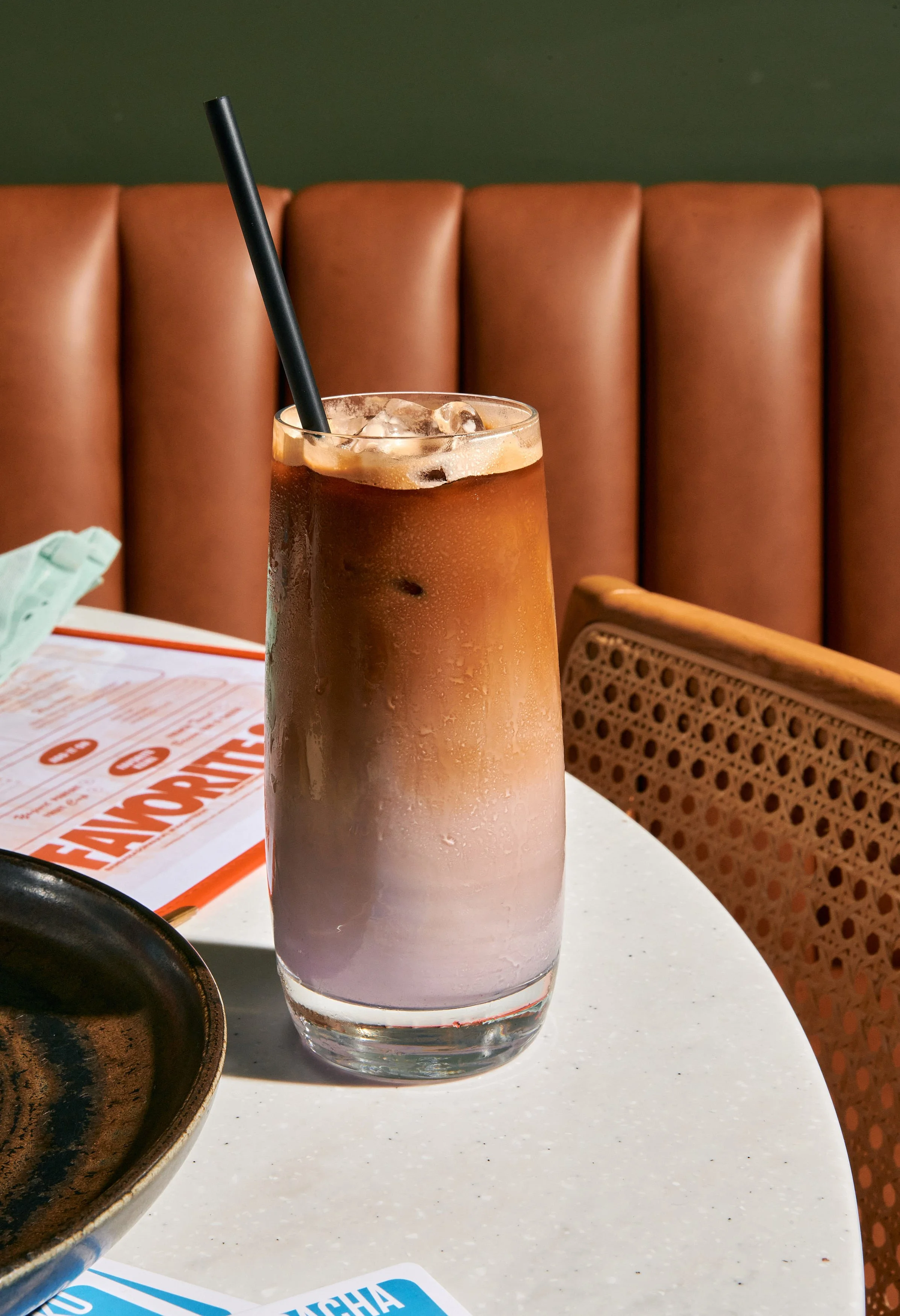 A tall glass of iced brown beverage with a straw, condensation on the outside, sitting on a white table in a restaurant setting with brown cushioned seats and a green wall in the background.