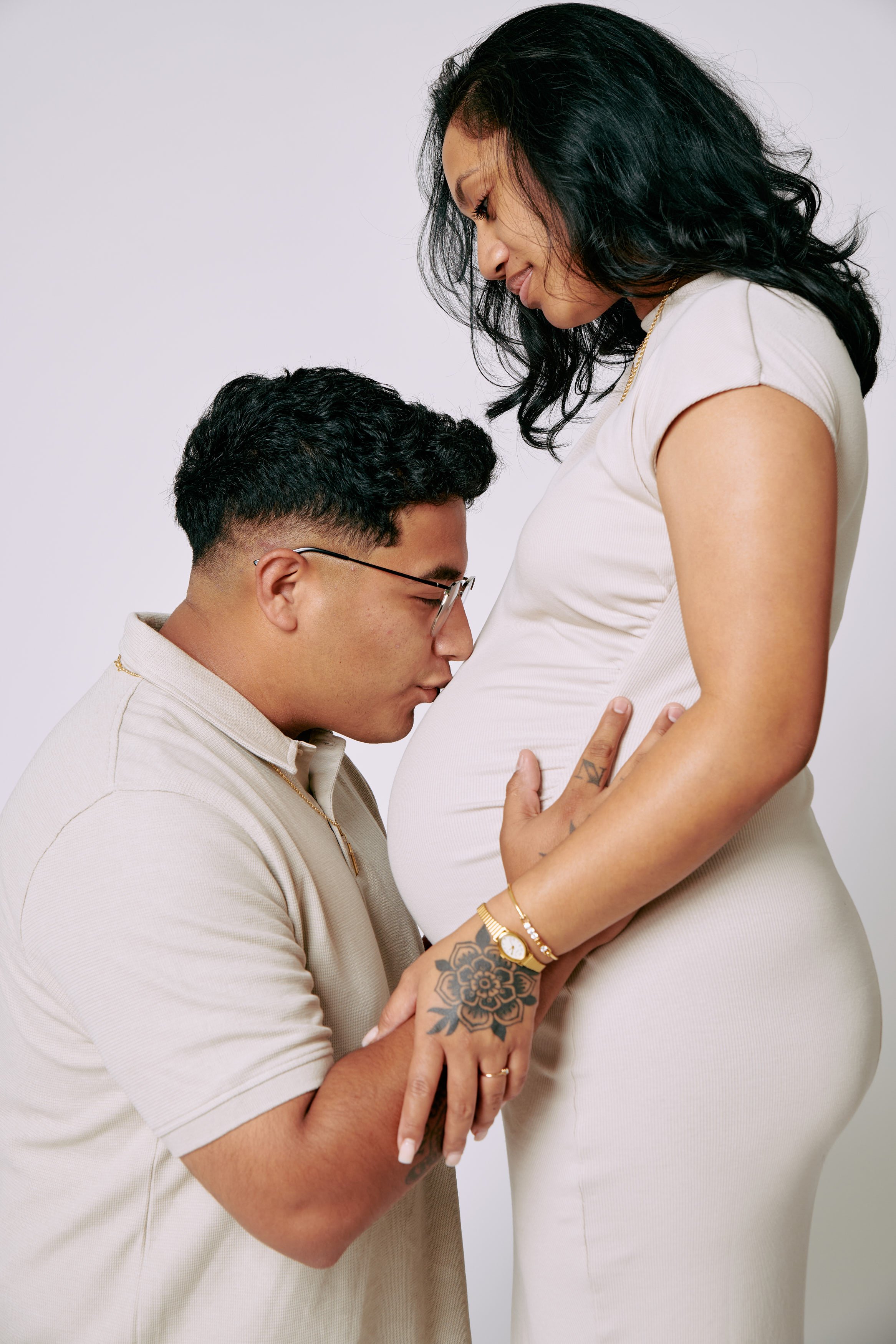 A man kneeling and kissing a pregnant woman's belly, with both looking at her stomach. The woman is standing, wearing a light-colored dress, and the man is wearing glasses and a light shirt.