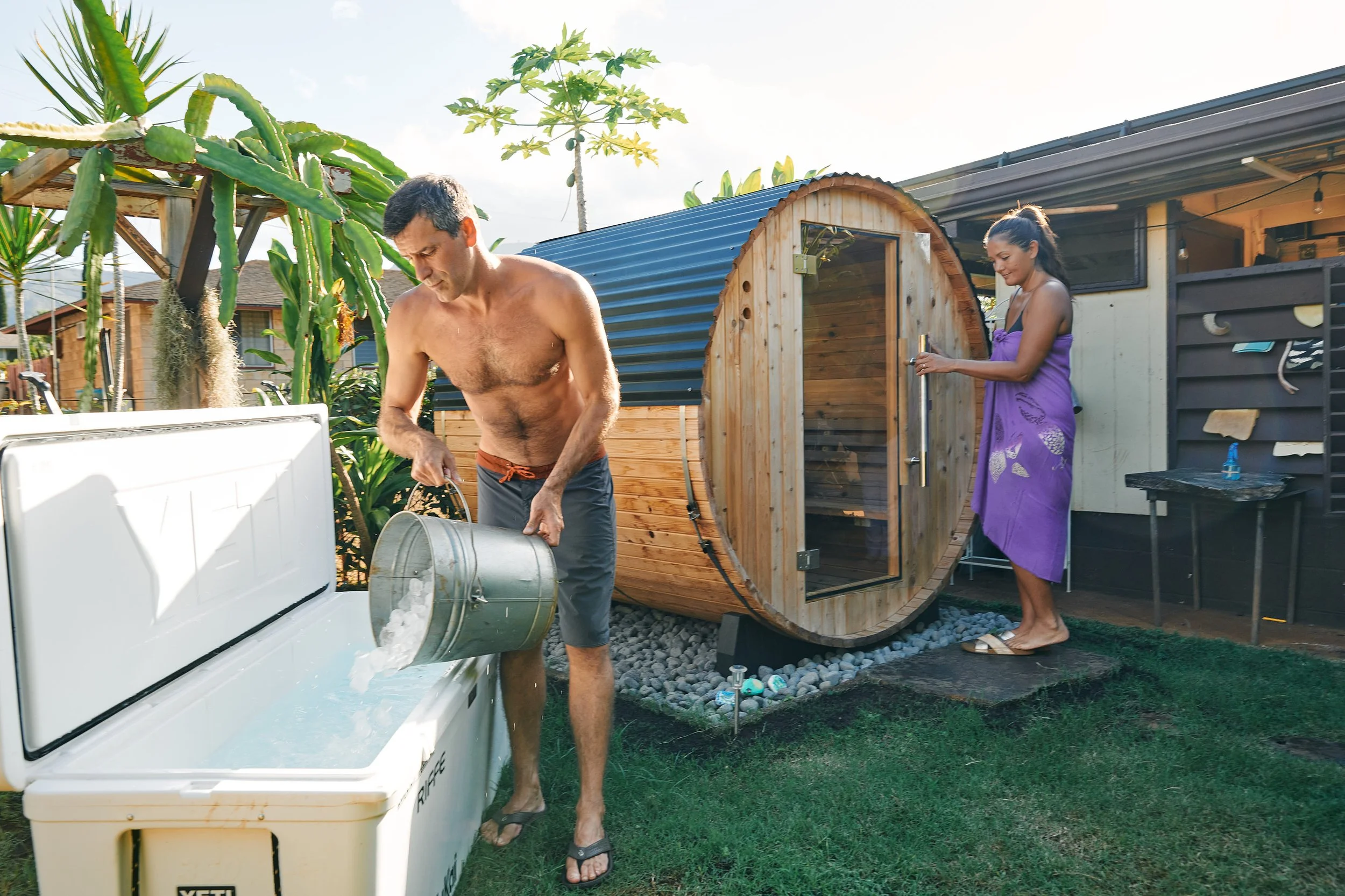 A man in swim trunks pouring ice into a cooler, a woman in a purple towel standing by a wooden sauna, in a backyard with tropical plants and a small shed.