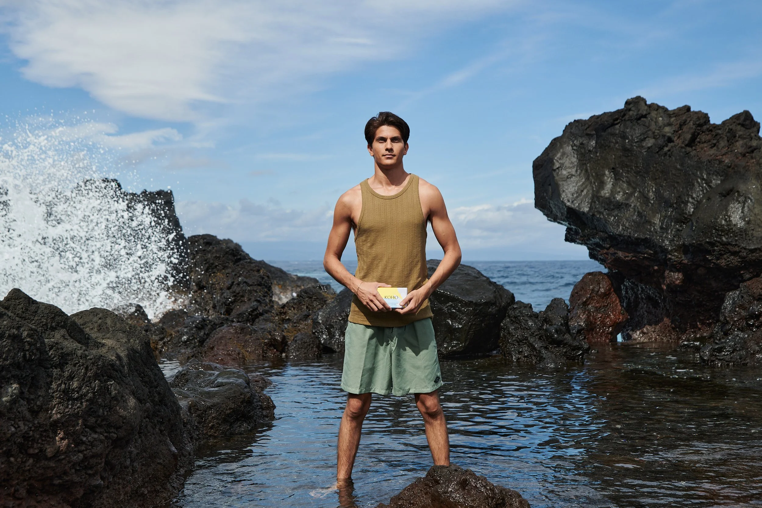 A young man standing in shallow water among black volcanic rocks on a coast, holding a box labeled 'KOHO', with an ocean and blue sky in the background.
