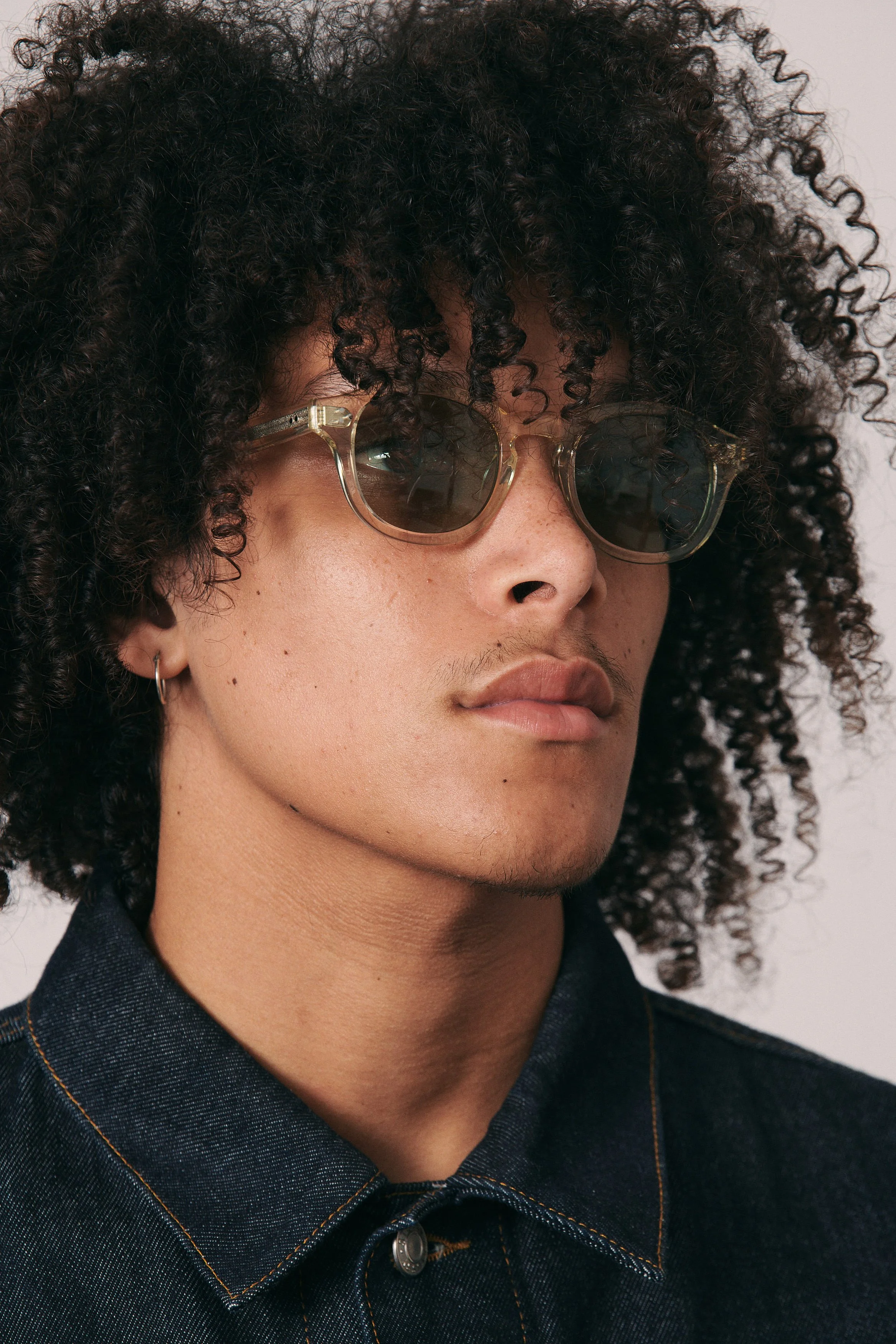 A person with curly hair wearing sunglasses and a dark denim shirt.