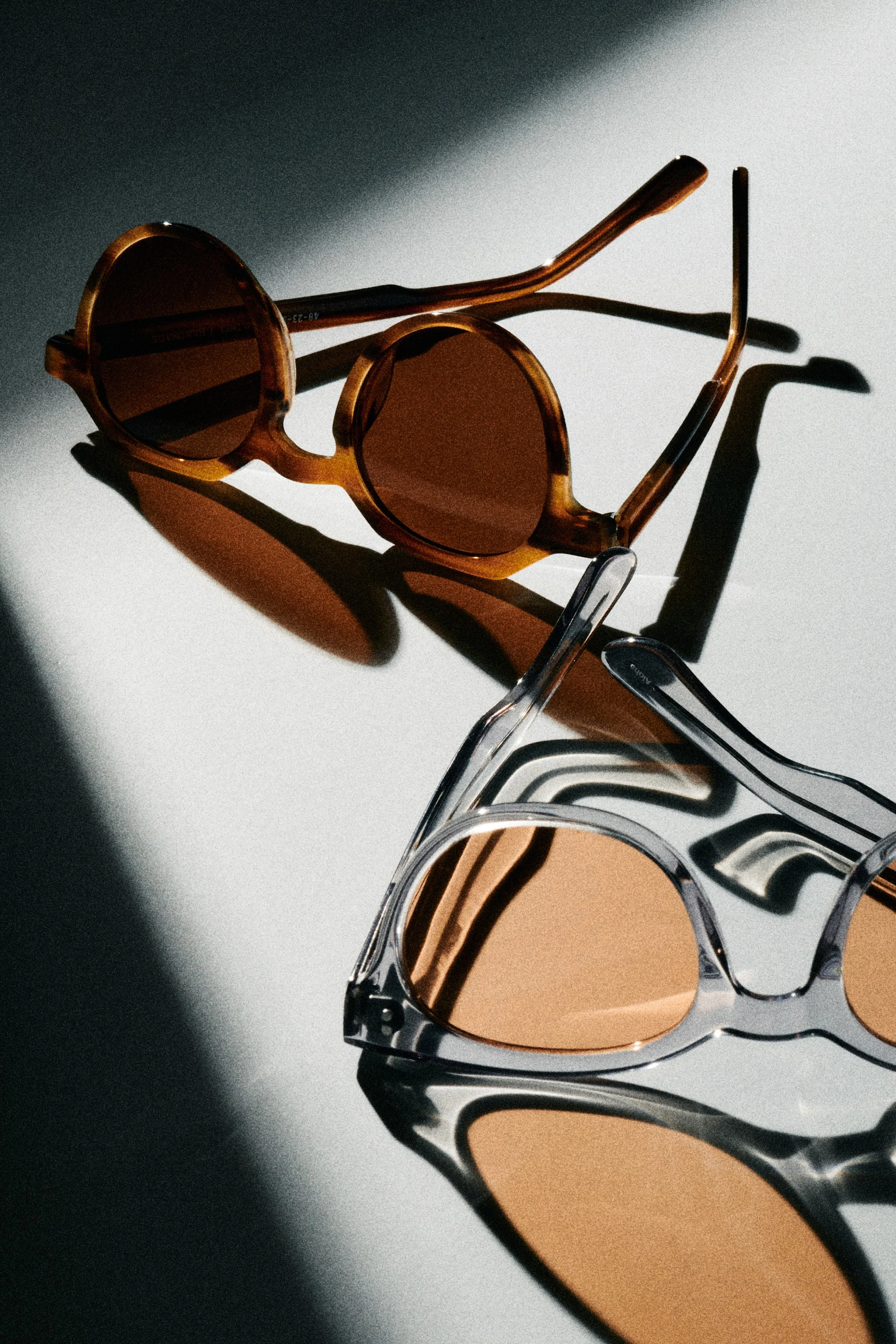 Two pairs of sunglasses, one with brown tortoiseshell frames and dark lenses, and the other clear frames with amber lenses, on a white surface with shadows cast by light source.