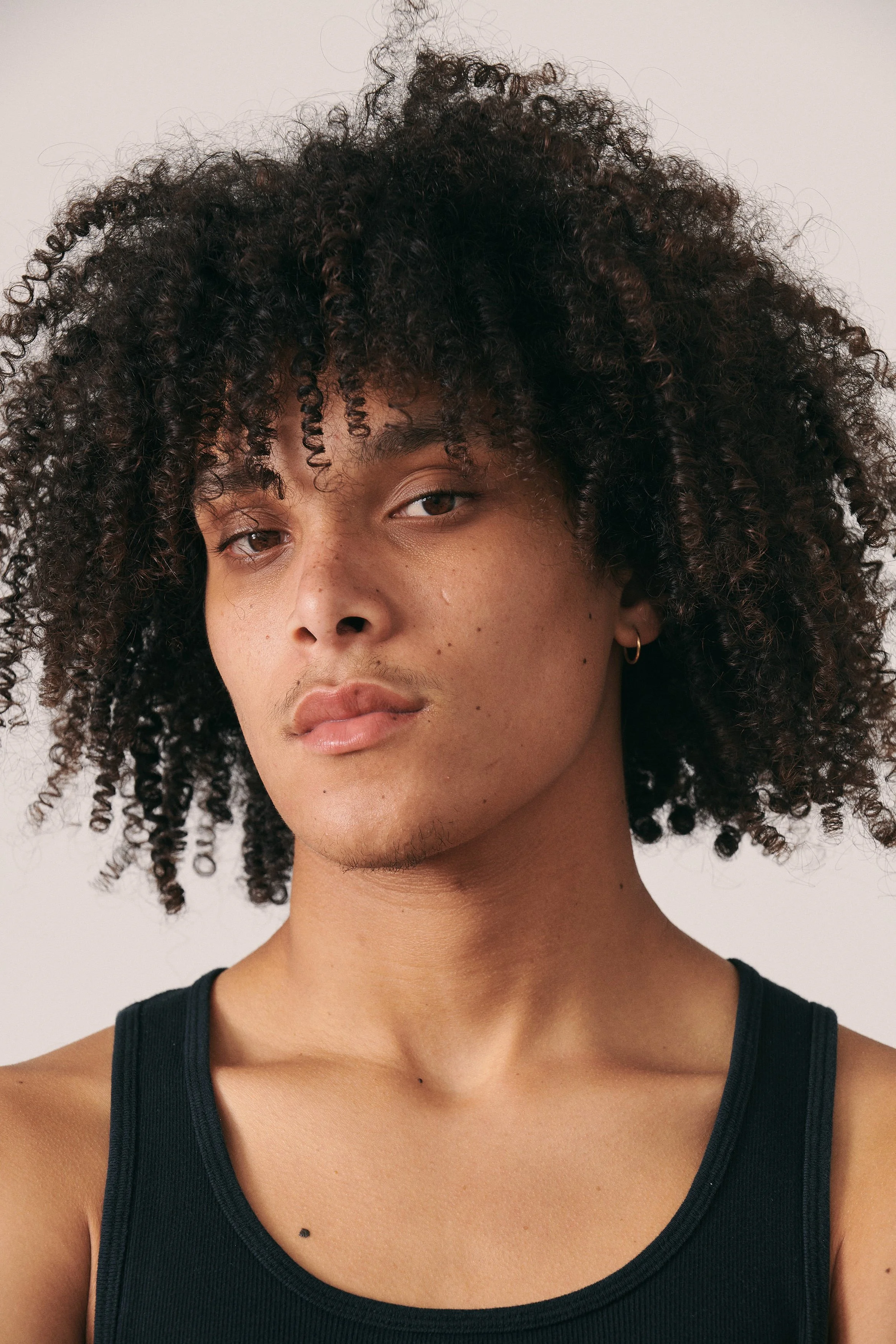 A young person with medium brown skin and curly dark hair, wearing a black tank top and a small hoop earring, looking directly at the camera with a neutral expression.