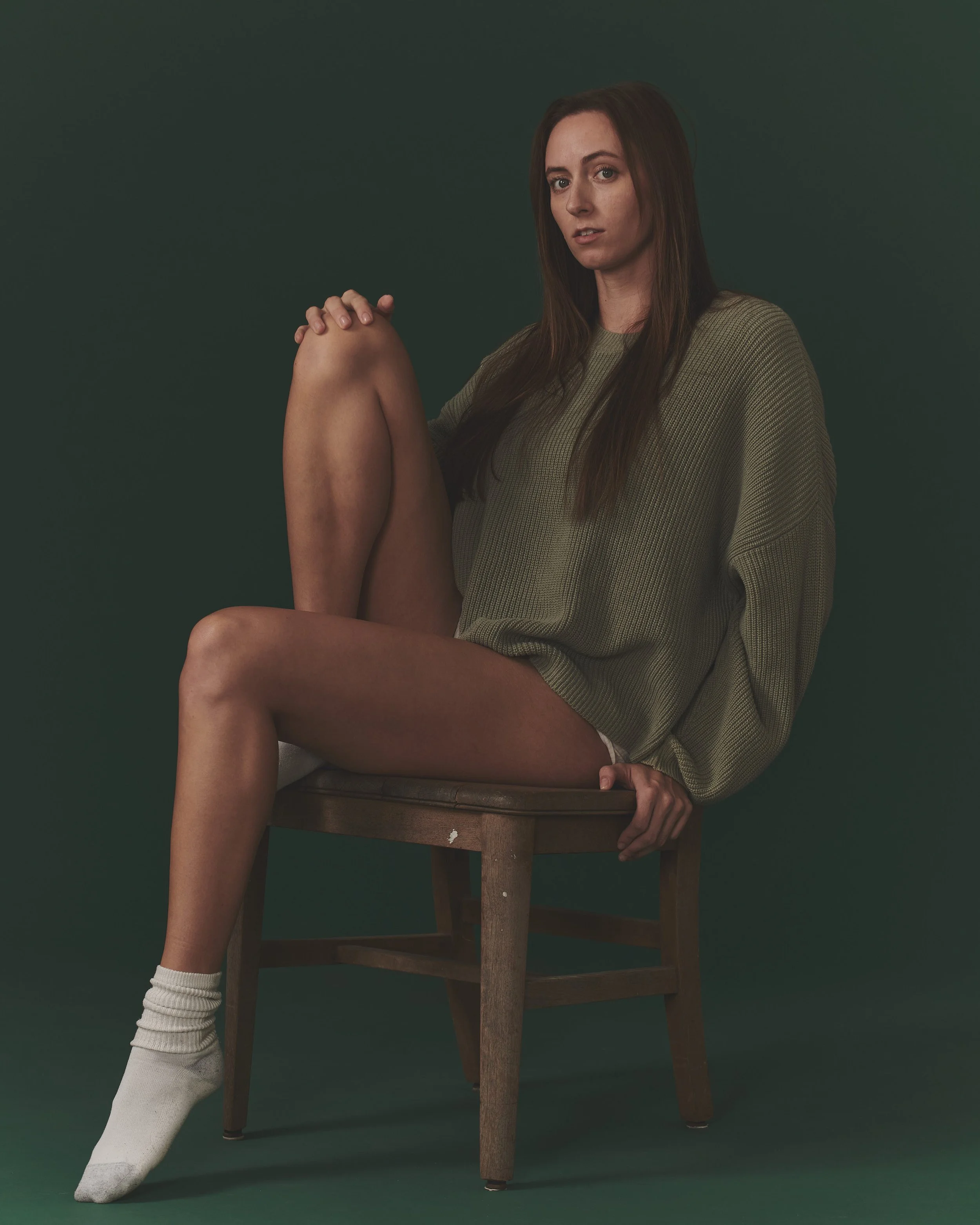 A woman with long brown hair is sitting on a wooden chair against a dark green background. She is wearing a loose olive green sweater, white socks, and no visible pants, with one leg bent and resting on the chair.