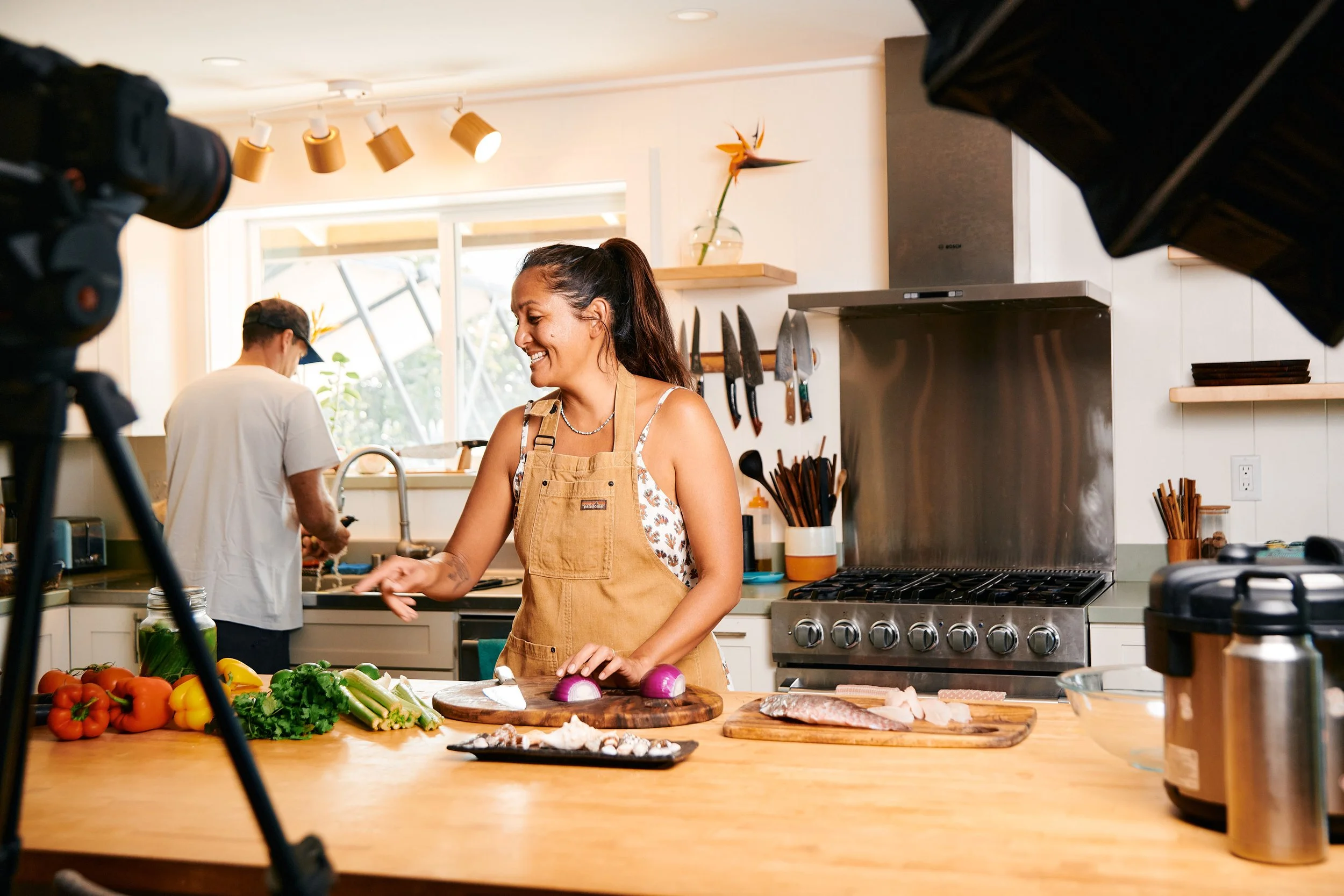 A woman cooking in a bright kitchen, chopping vegetables while filming a cooking show, with a man washing dishes in the background.