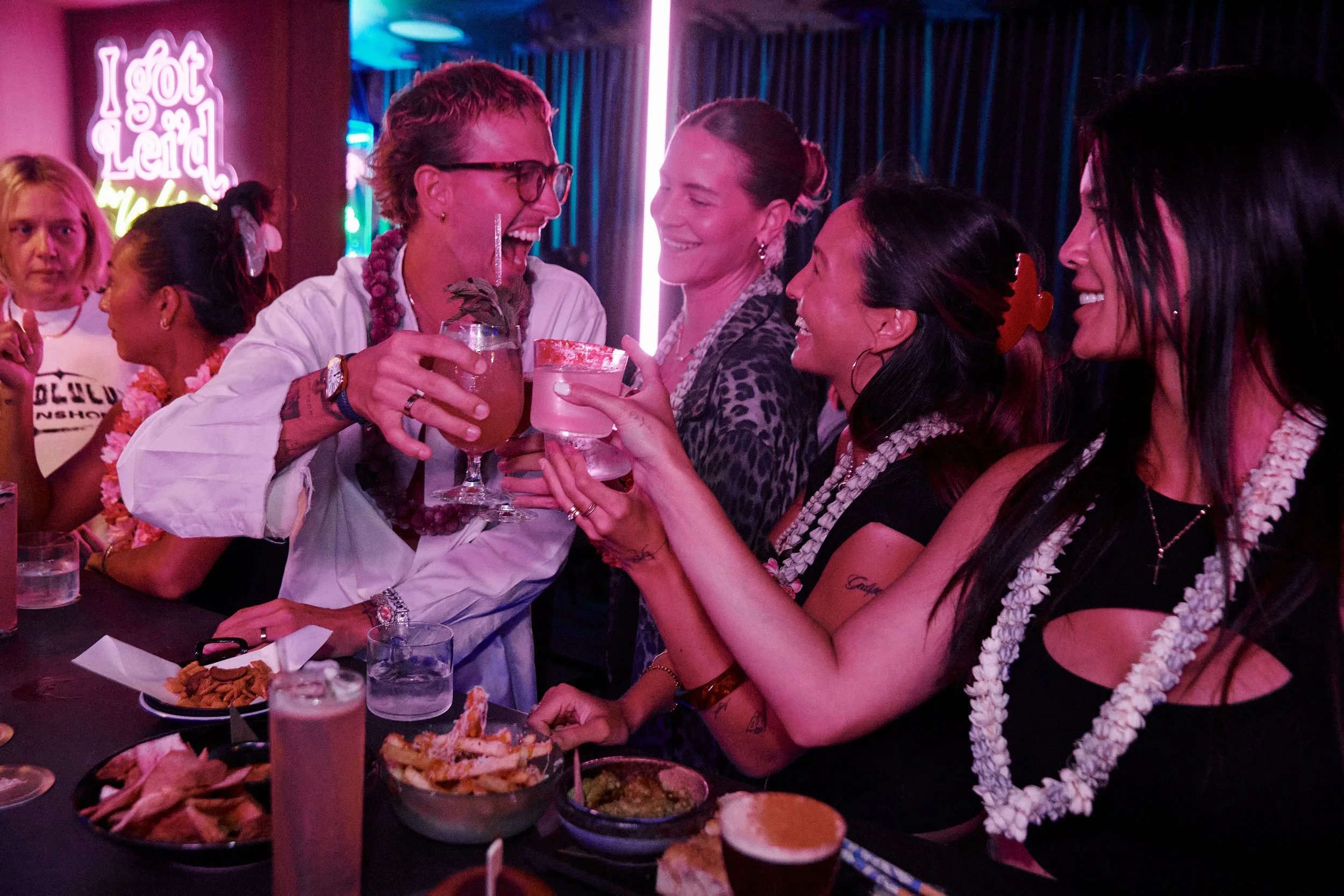 Group of women celebrating at a bar with drinks and snacks, in a lively setting with neon signs and colorful lighting.