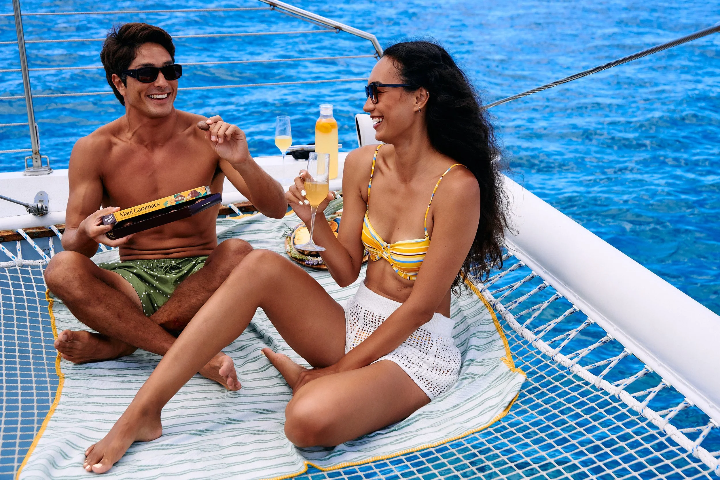 A man and woman sitting on a boat's netting, enjoying drinks and smiling at each other, with the ocean in the background.