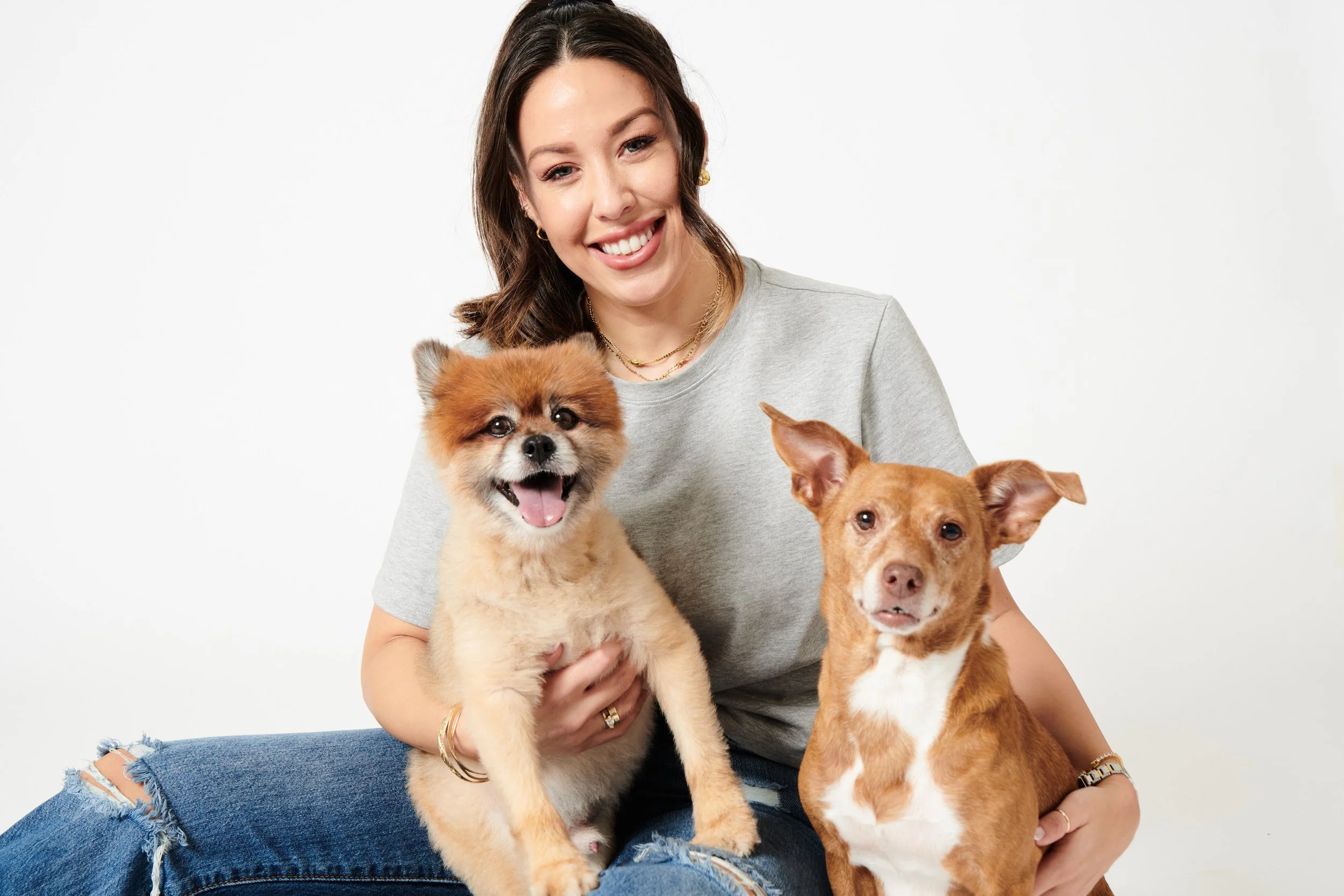A woman smiling and holding two small dogs, one with fluffy fur and the other with short fur, against a plain white background.