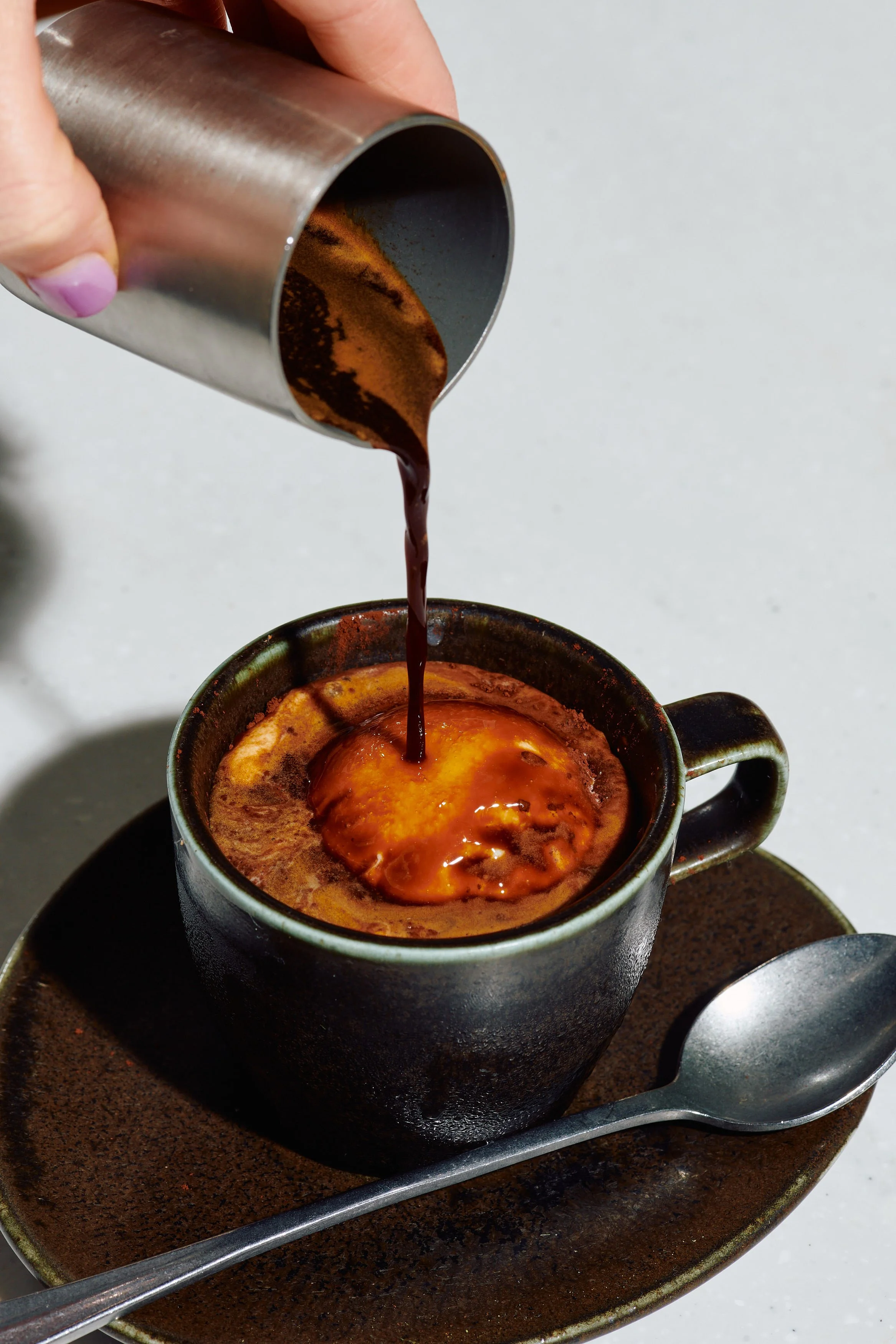 Hot coffee in a black mug with steam rising, chocolate syrup being poured into the mug from a metal container, with a silver spoon on a matching saucer.