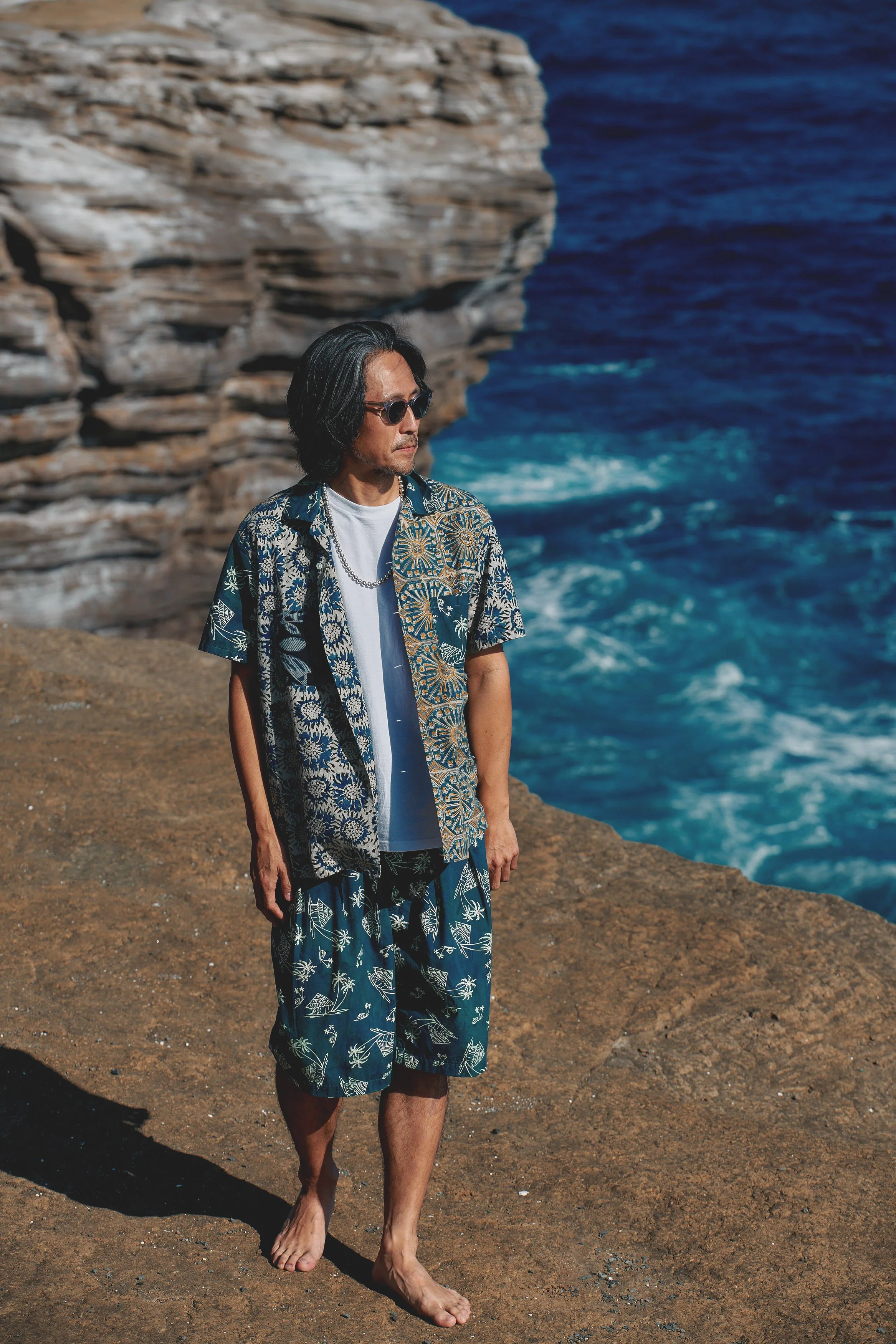 A man walking barefoot on a rocky coastline, wearing sunglasses, a patterned shirt, shorts, and a chain necklace, with the ocean and rocky cliffs in the background.