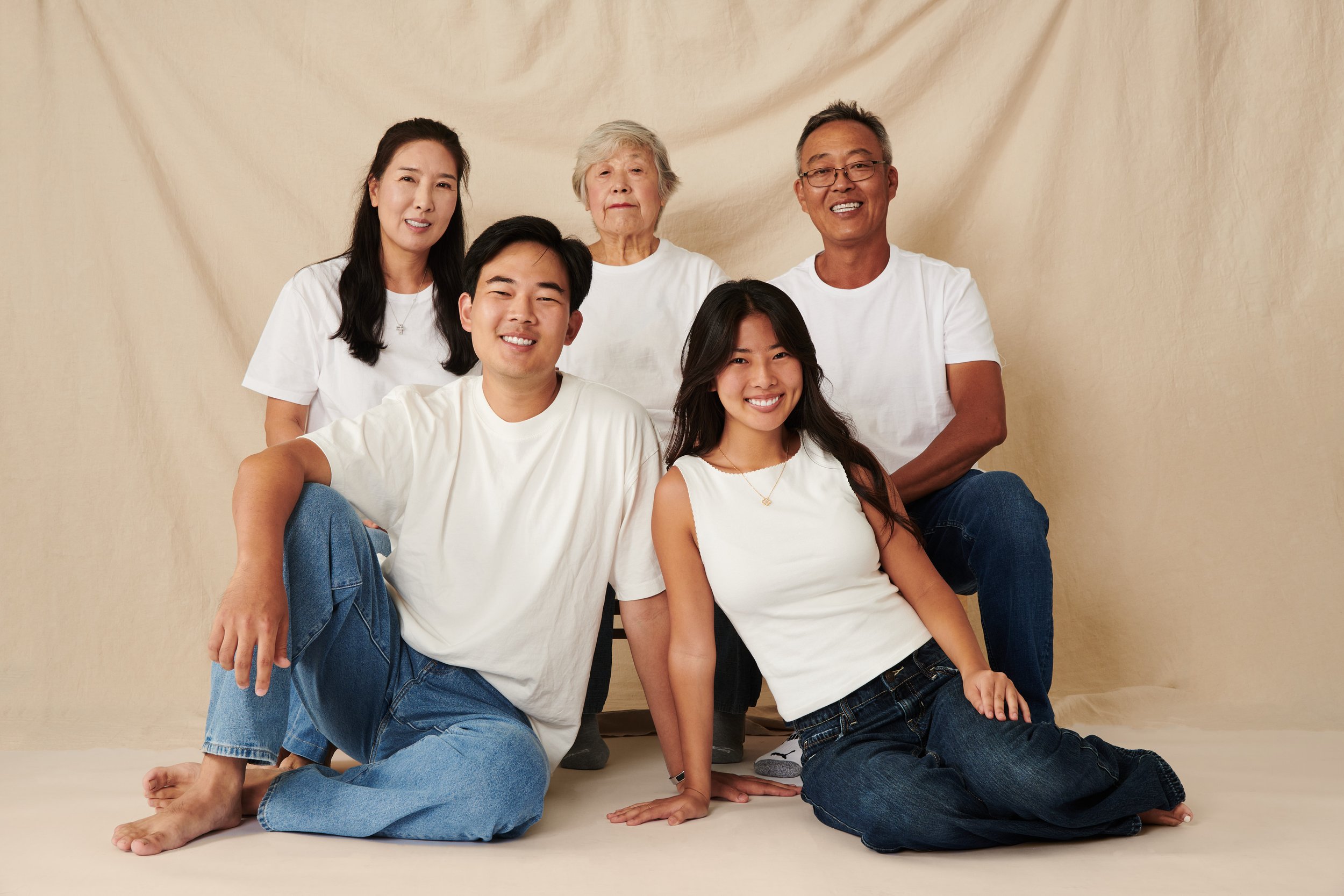 Group of six diverse family members posing together, sitting and kneeling on the floor, against a plain beige backdrop.