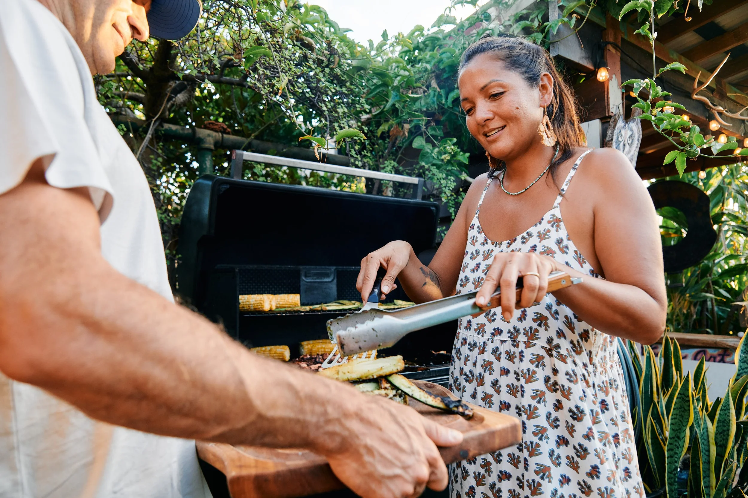 Two people grilling vegetables, such as zucchini and corn, outdoors with a grill in the background, surrounded by greenery and string lights.