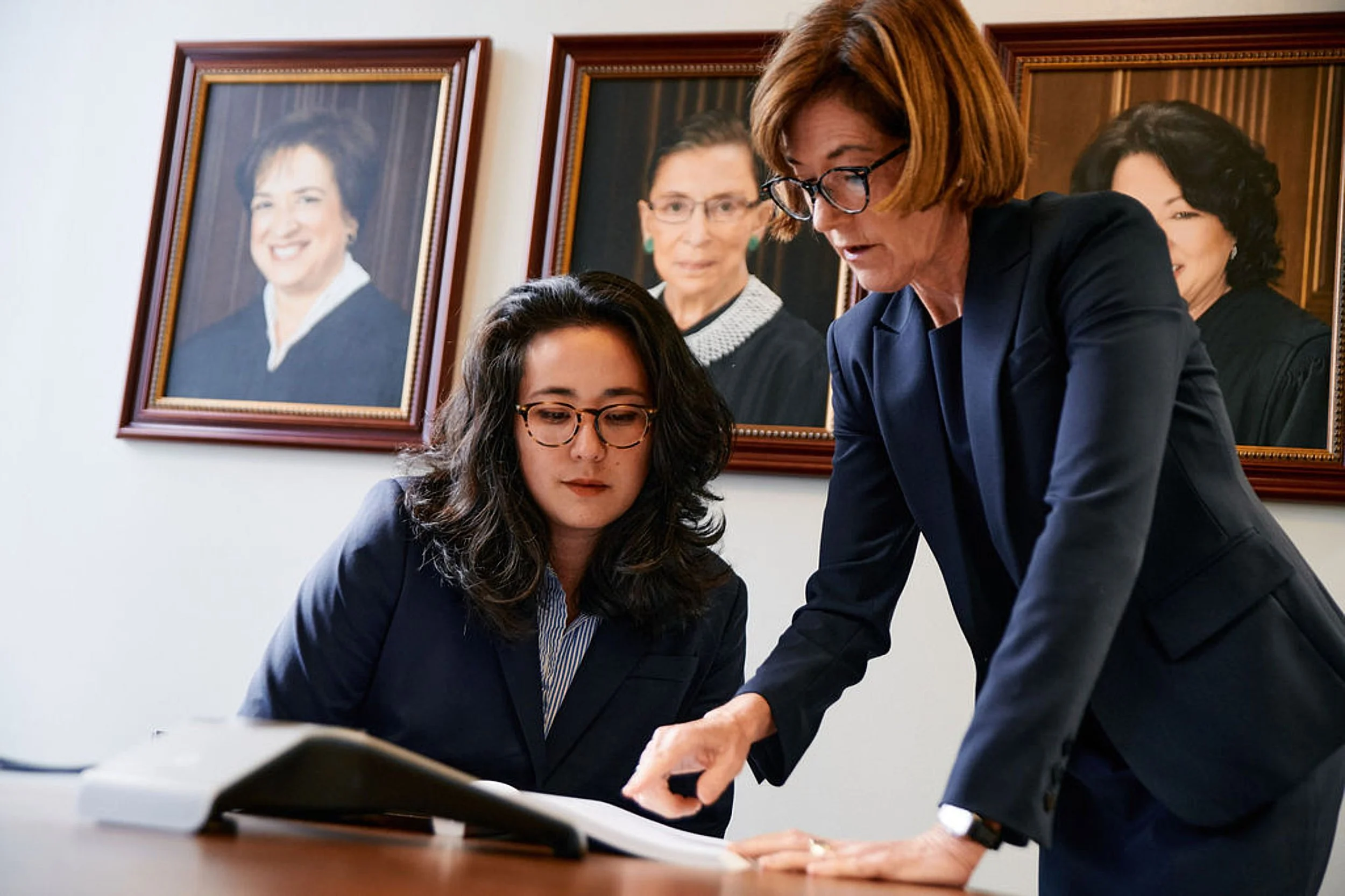 Two professional women reviewing documents in an office, with portrait photographs of women in judicial robes hanging on the wall behind them.