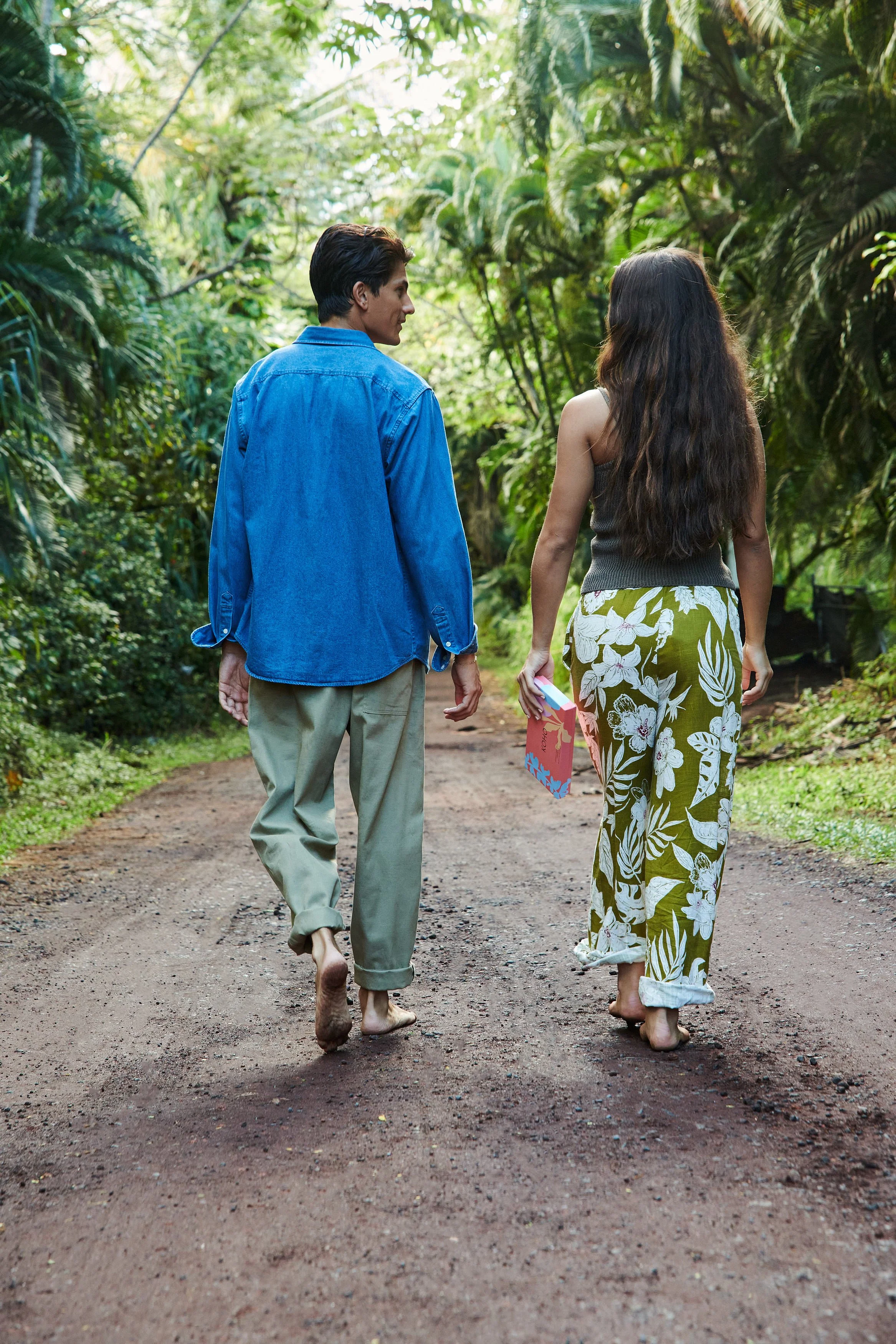 A man and woman walking barefoot on a dirt path through a lush green jungle, holding hands and looking at each other.