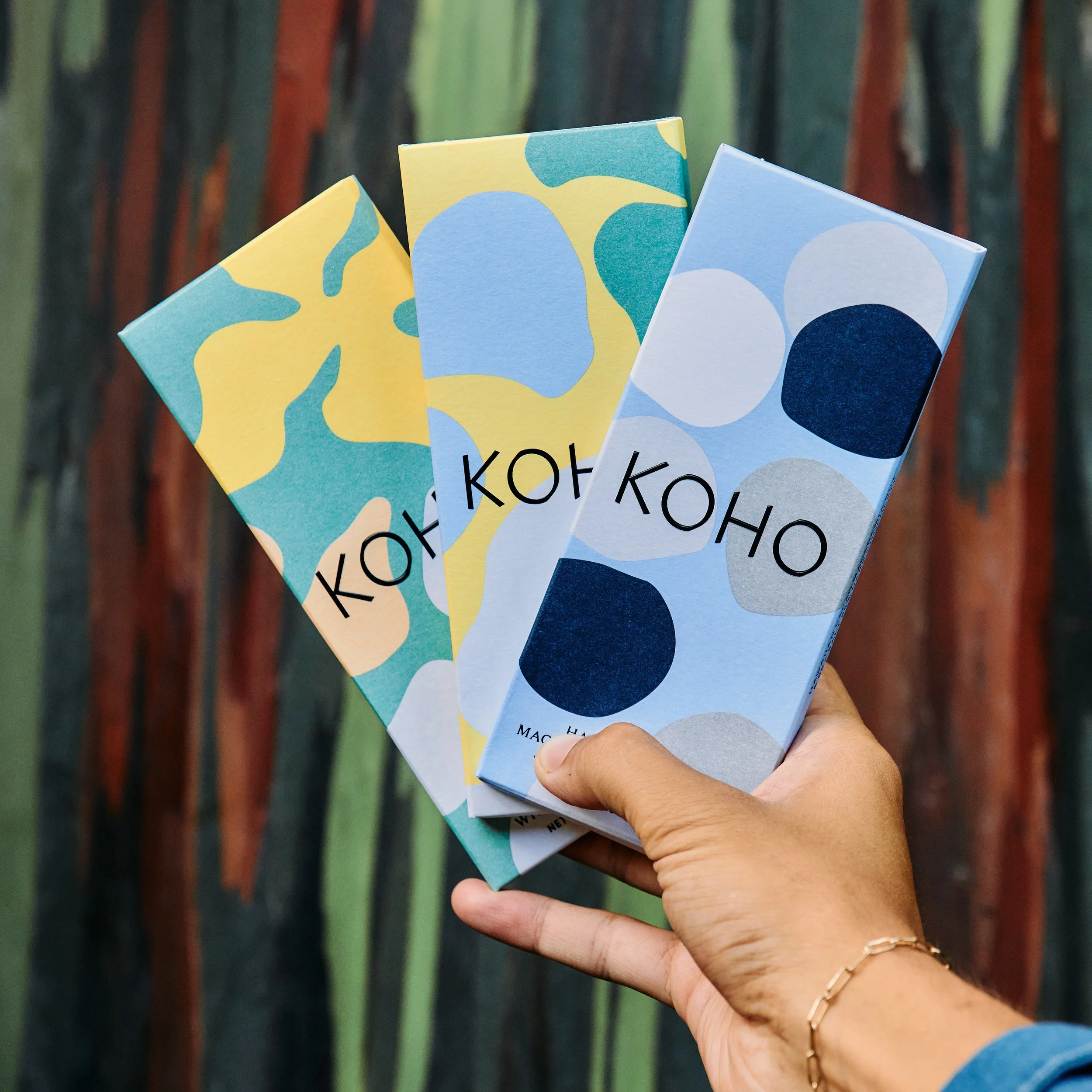 Hand holding three Kozoko brand colorful abstract patterned boxes with a wooden fence in the background.