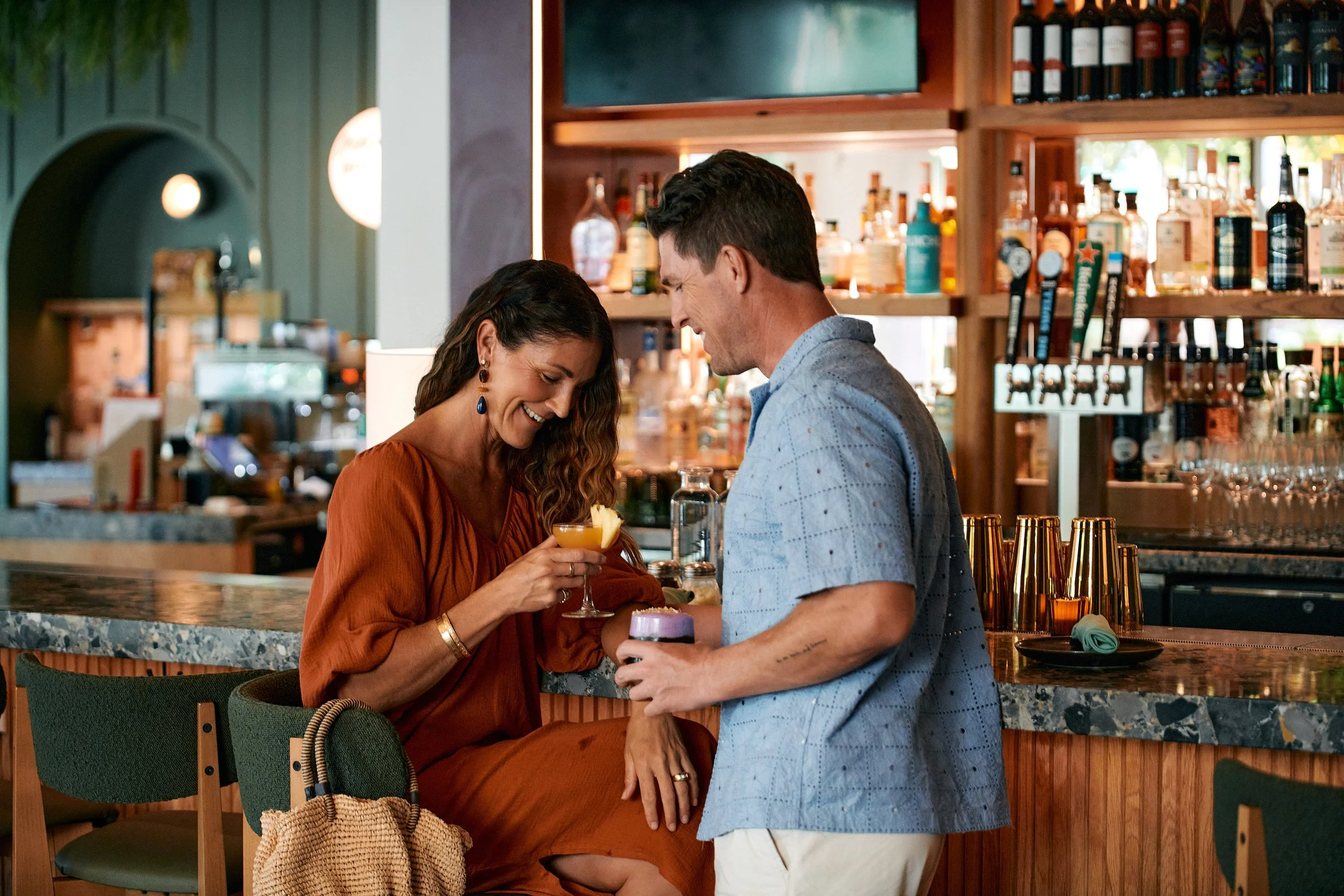 A woman and man sharing a laugh at a bar, with the woman holding a cocktail and the man holding a drink. The bar has liquor bottles on shelves behind them.