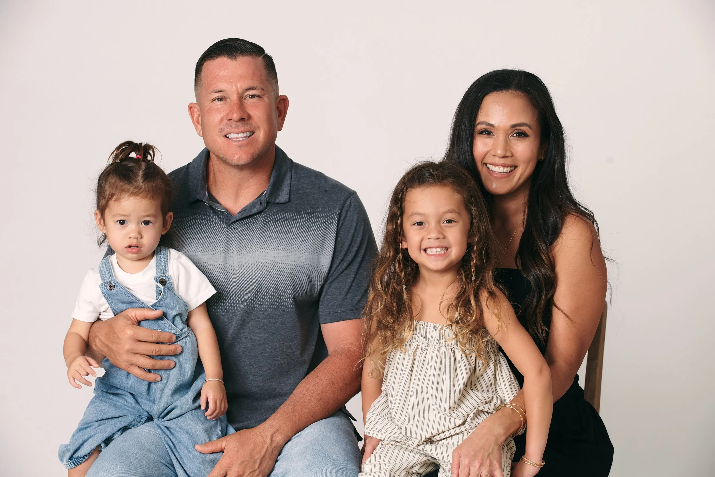 A happy family of four, including a man, a woman, and two young girls, posing together against a plain white background.
