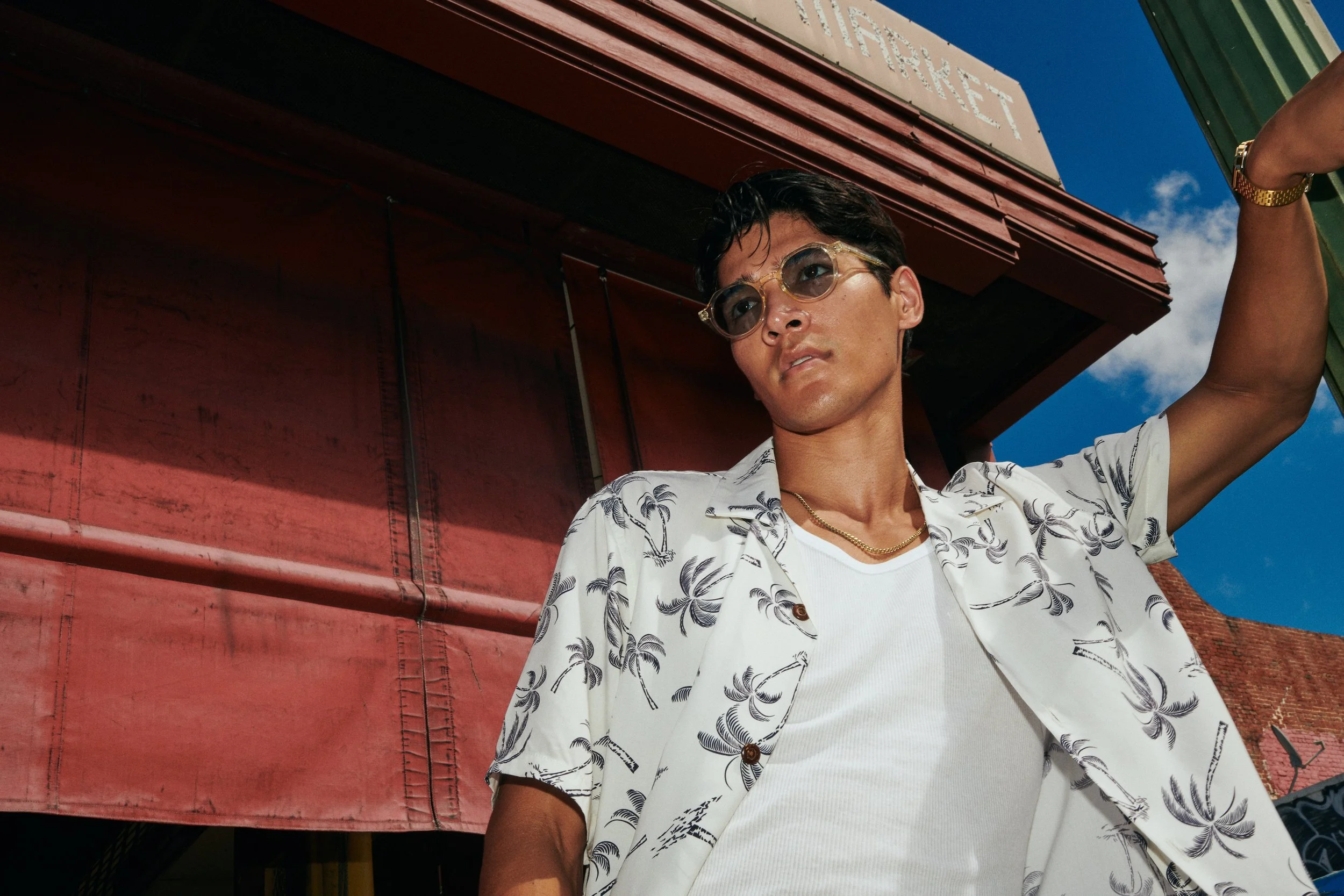 Young man wearing sunglasses and a floral shirt, standing outdoors with a red building and a blue sky in the background.
