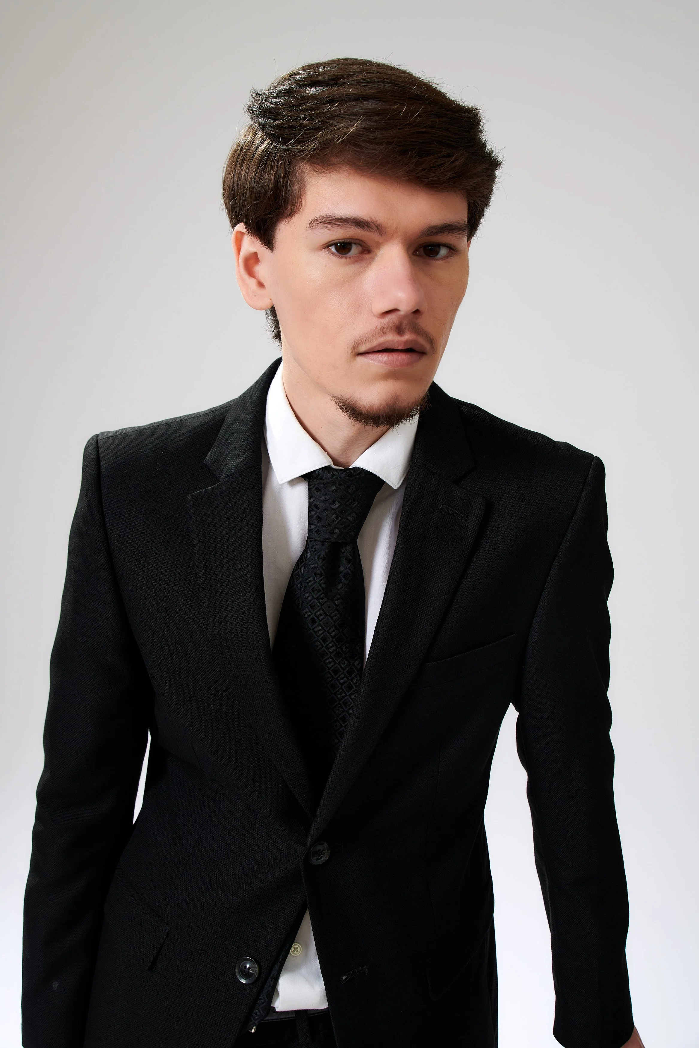 A young man with brown hair, light facial hair, wearing a black suit, white shirt, and black tie. He has a serious expression and is standing against a plain light background.