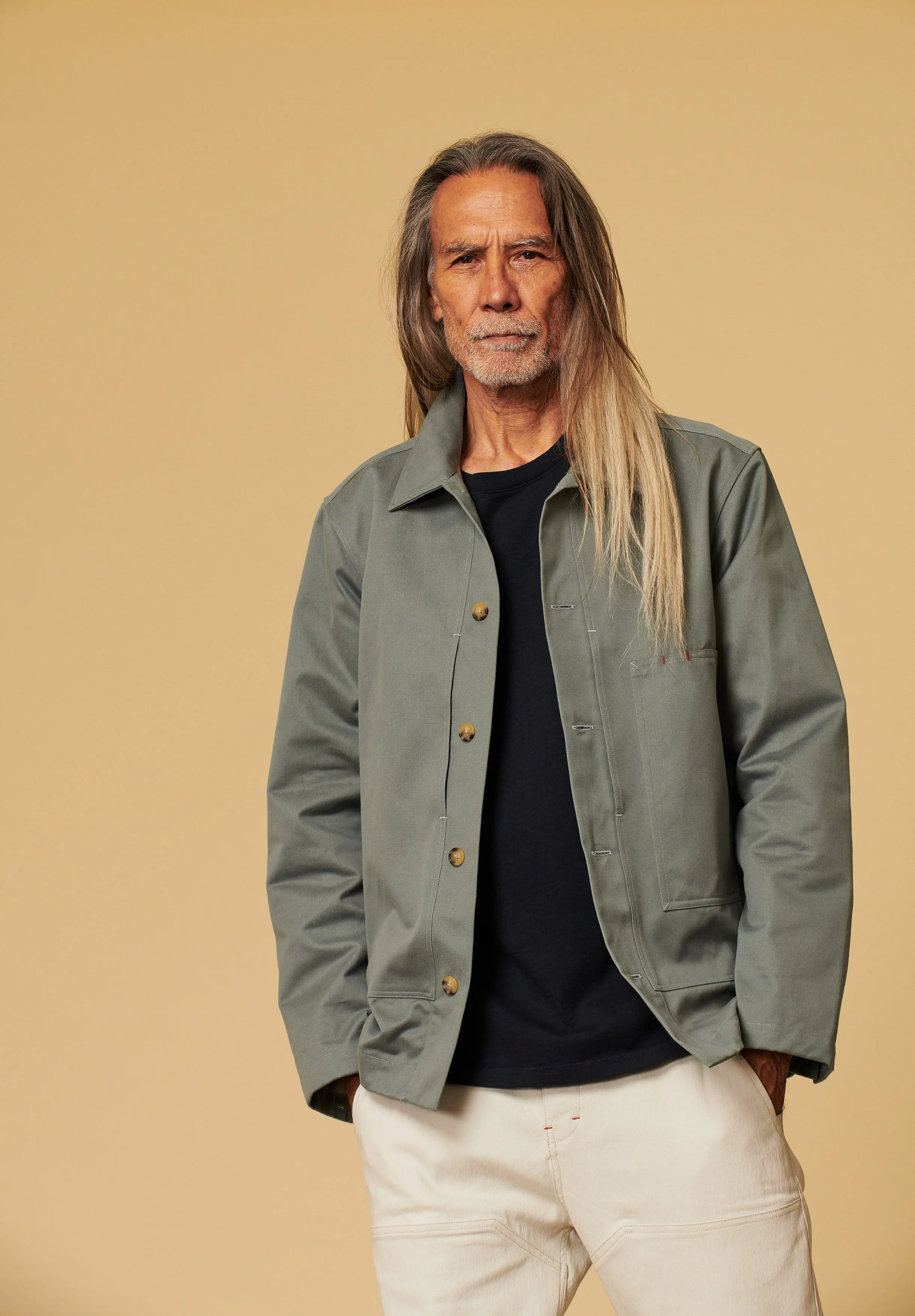 Older man with long hair and beard, wearing a gray jacket over a black shirt and beige pants, standing against a plain yellow background.