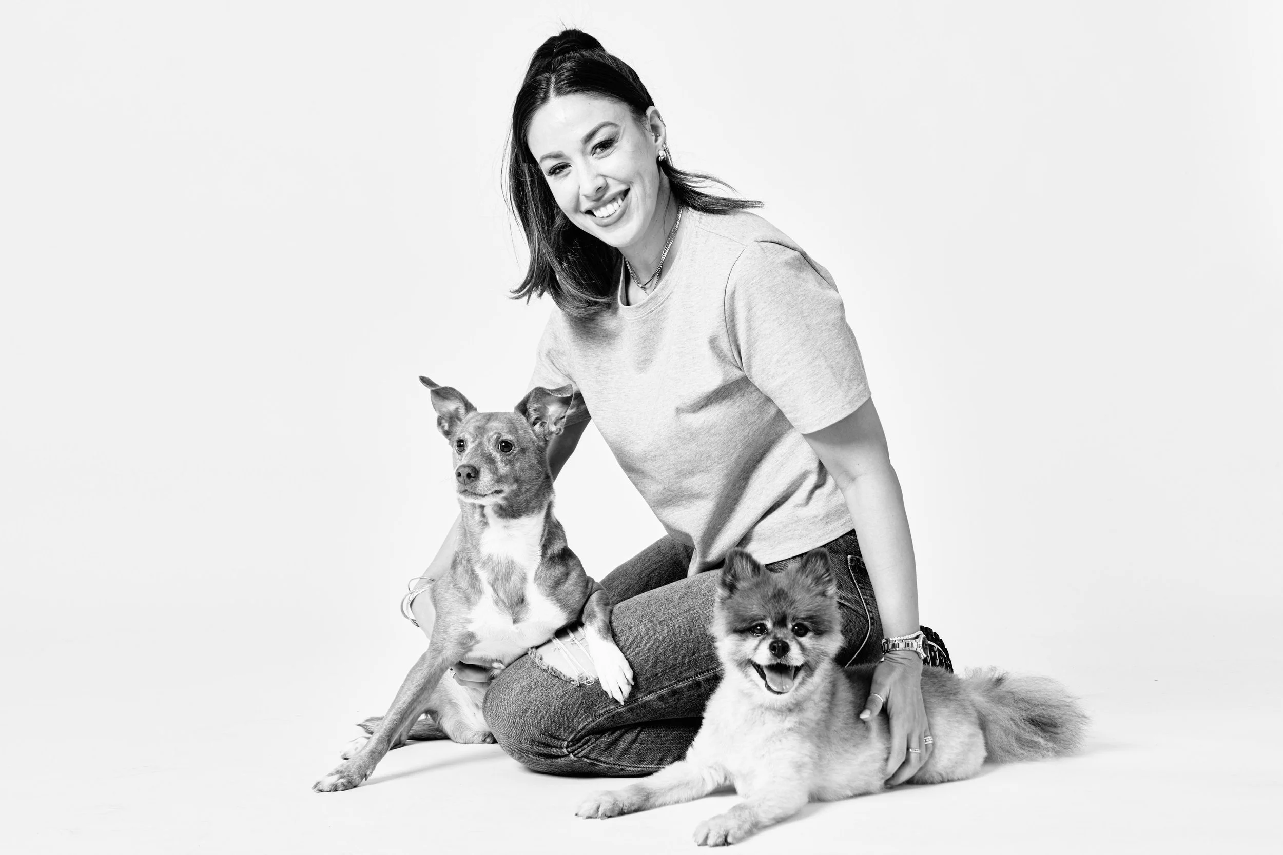 A woman sitting on the floor with two dogs, one puppy with a fluffy coat and a smile, and another slightly older dog with a lean body, both dogs sitting close to her.